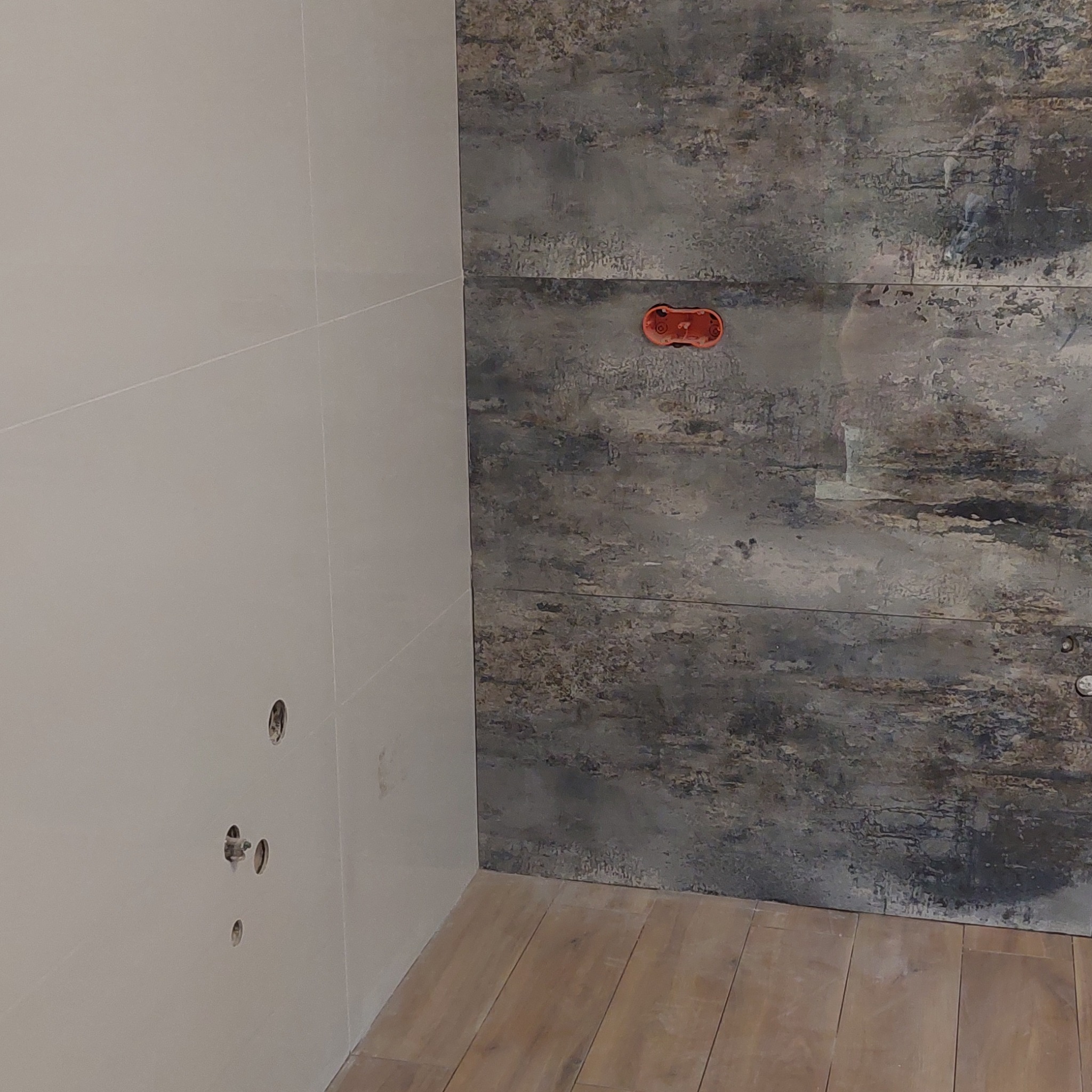 Fragment ściany z dwiema różnymi okładzinami: jasne płytki i ciemne panele dekoracyjne imitujące kamień, widoczne otwory na instalacje elektryczne i drewnopodobna podłoga.