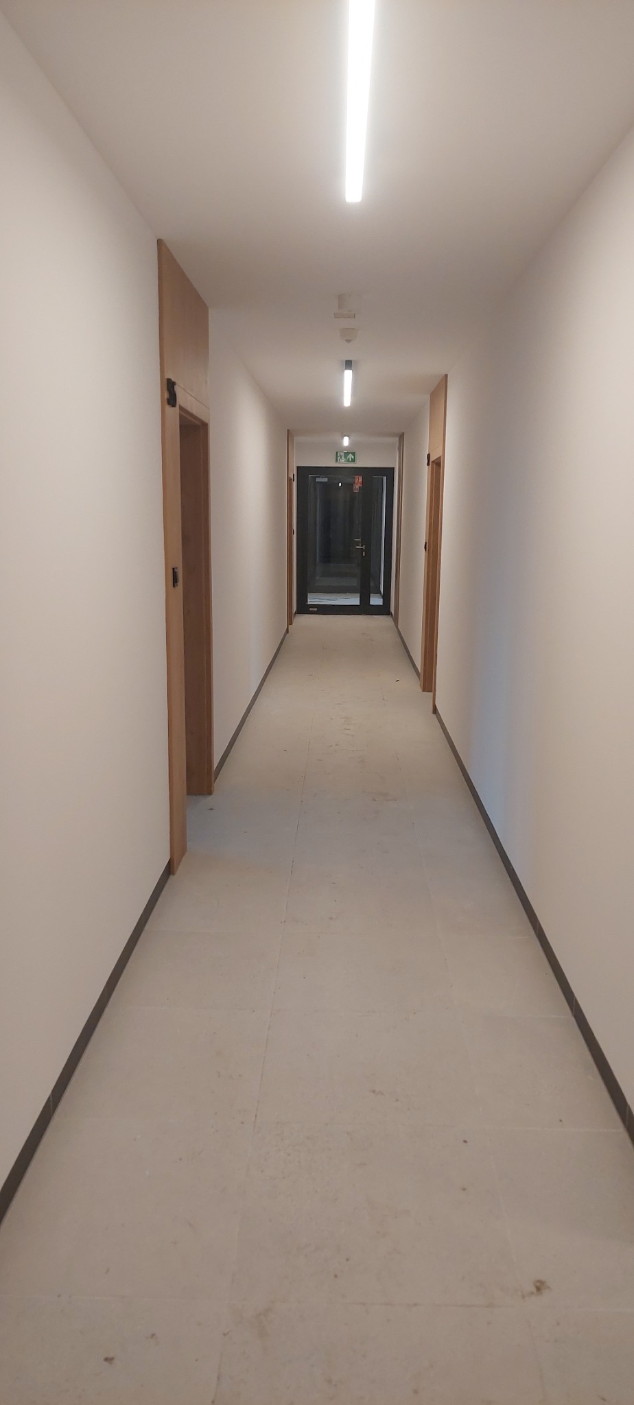 Długi korytarz w nowym budynku z białymi ścianami, jasną podłogą i drewnianymi drzwiami po bokach, zakończony czarnymi drzwiami ewakuacyjnymi.