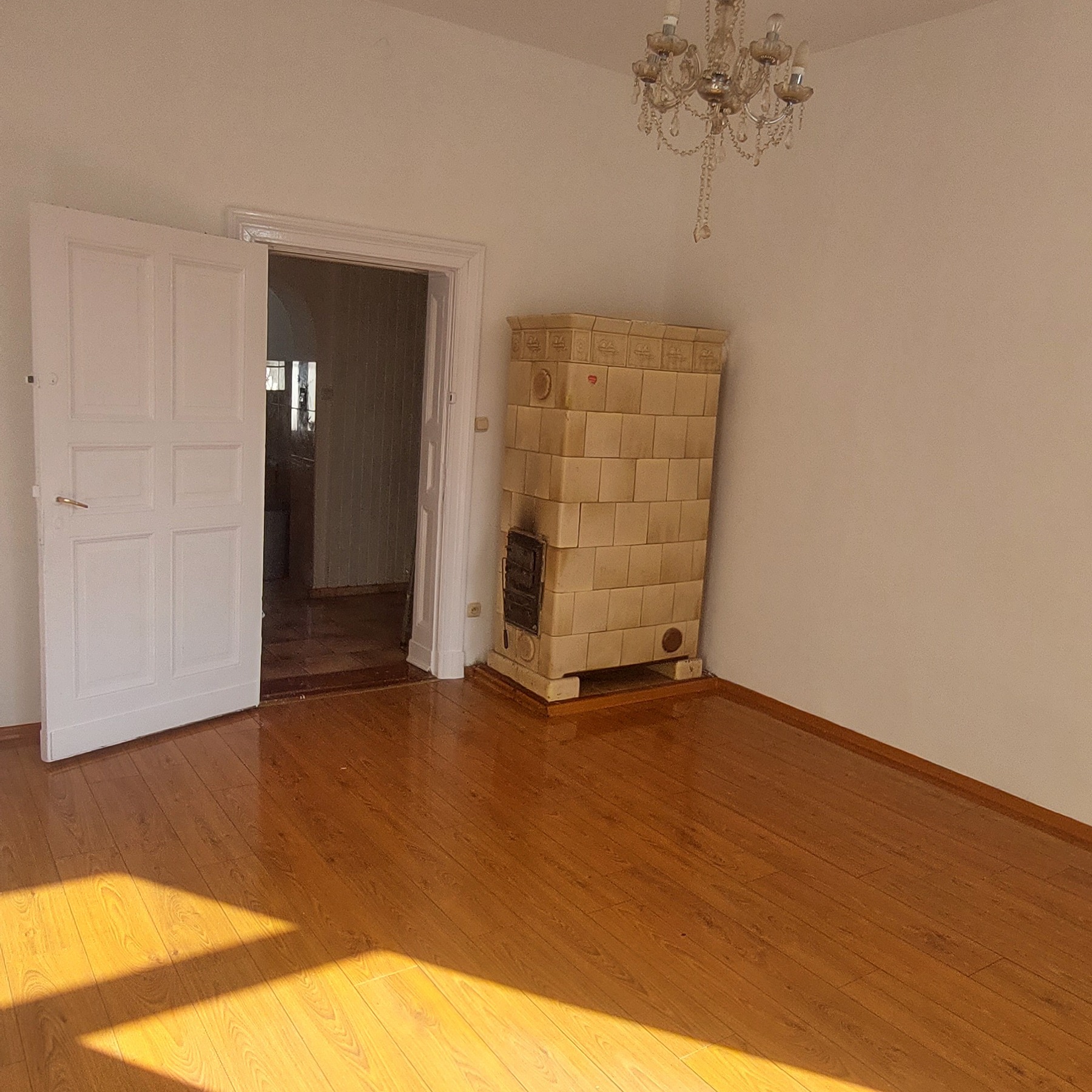 Pokój z drewnianą podłogą, otwartymi białymi drzwiami, kaflowym piecem i kryształowym żyrandolem