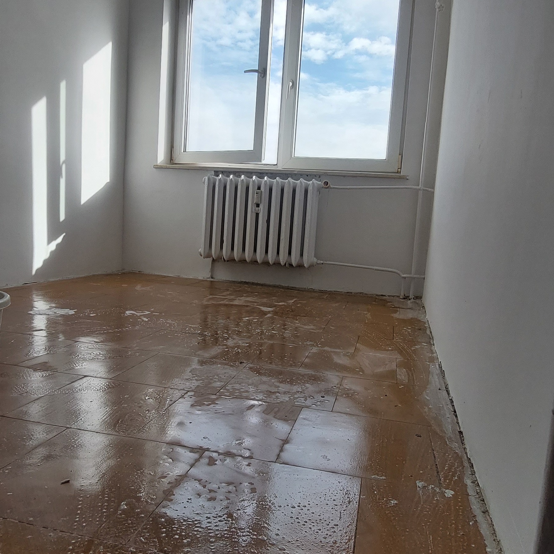 Pokój z mokrą, brązową podłogą z płytek, prawdopodobnie po umyciu lub w trakcie czyszczenia. Widoczne białe ściany, okno z widokiem na niebo i grzejnik.