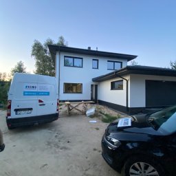 PPHU Miłosz Weistock - Nowoczesny, dwukondygnacyjny dom z białymi ścianami i czarnymi ramami okiennymi po zakończeniu budowy, z widocznym samochodem osobowym i dostawczym firmy sprzątającej. Na masce samochodu osobowego...