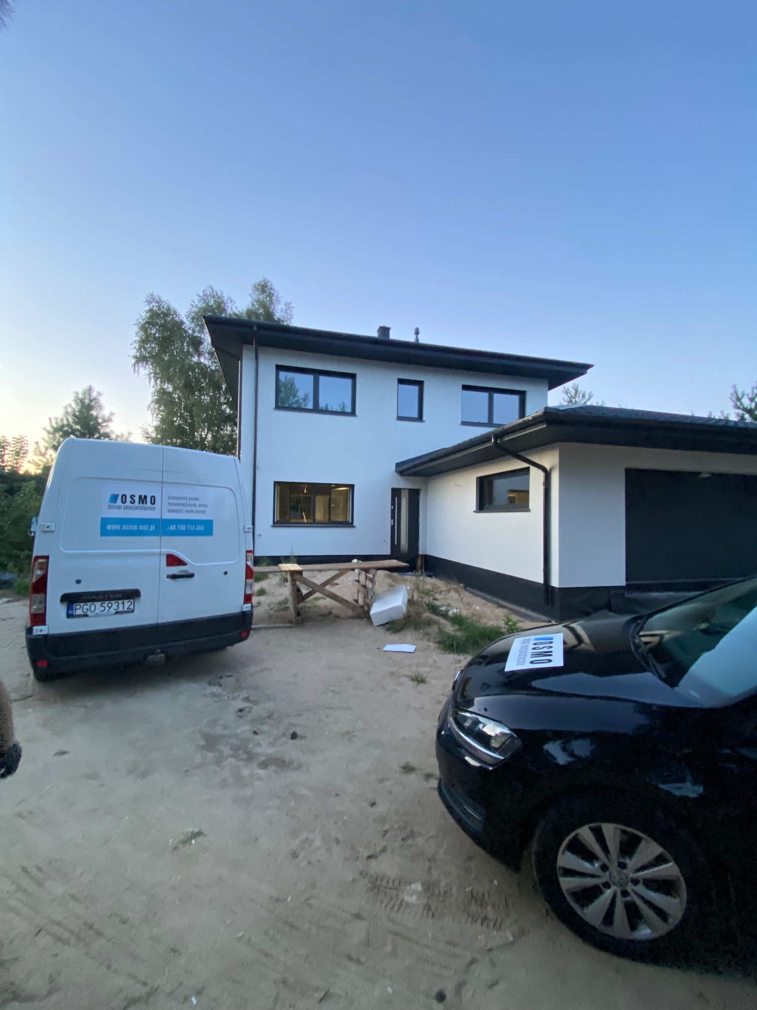 Nowoczesny, dwukondygnacyjny dom z białymi ścianami i czarnymi ramami okiennymi po zakończeniu budowy, z widocznym samochodem osobowym i dostawczym firmy sprzątającej. Na masce samochodu osobowego...