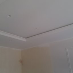 DOM SERWIS Krzysztof Chodorowski - Wykończony sufit podwieszany z listwami dekoracyjnymi i punktowym oświetleniem, fragment ściany w odcieniach bieli i beżu z widoczną instalacją elektryczną.
