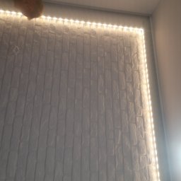 DOM SERWIS Krzysztof Chodorowski - Ściana z białej cegły dekoracyjnej oświetlona taśmą LED, widoczne niedoskonałości w ułożeniu cegieł i wystający fragment czyjejś głowy w rogu.