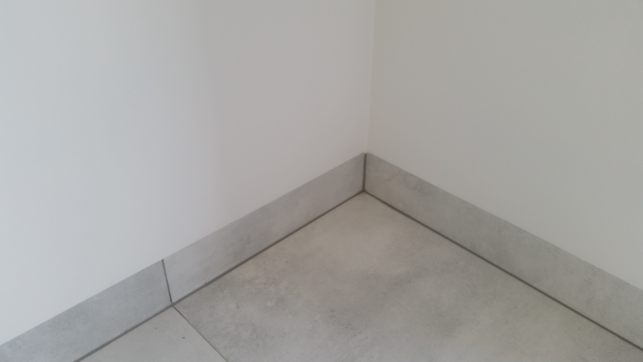 Szare płytki podłogowe i przypodłogowe w rogu pomieszczenia, minimalistyczny design, fuga w kolorze grafitowym.