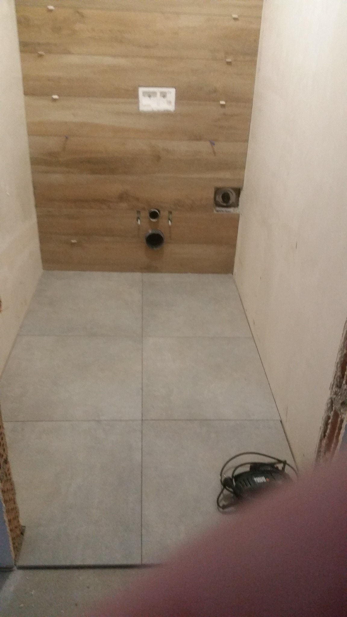 Wykończona podłoga z szarych płytek w łazience, ściana z drewnopodobnych paneli, widoczne otwory na instalację sanitarną, w rogu leży czarny kabel od elektronarzędzia.