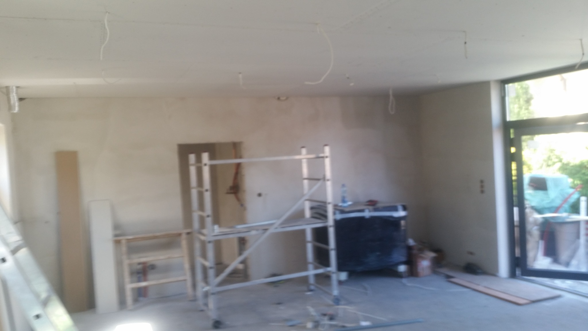 Wnętrze w trakcie prac wykończeniowych: ściany po szpachlowaniu, widoczne kable wystające z sufitu, aluminiowe rusztowanie i okno balkonowe z widokiem na zieleń.