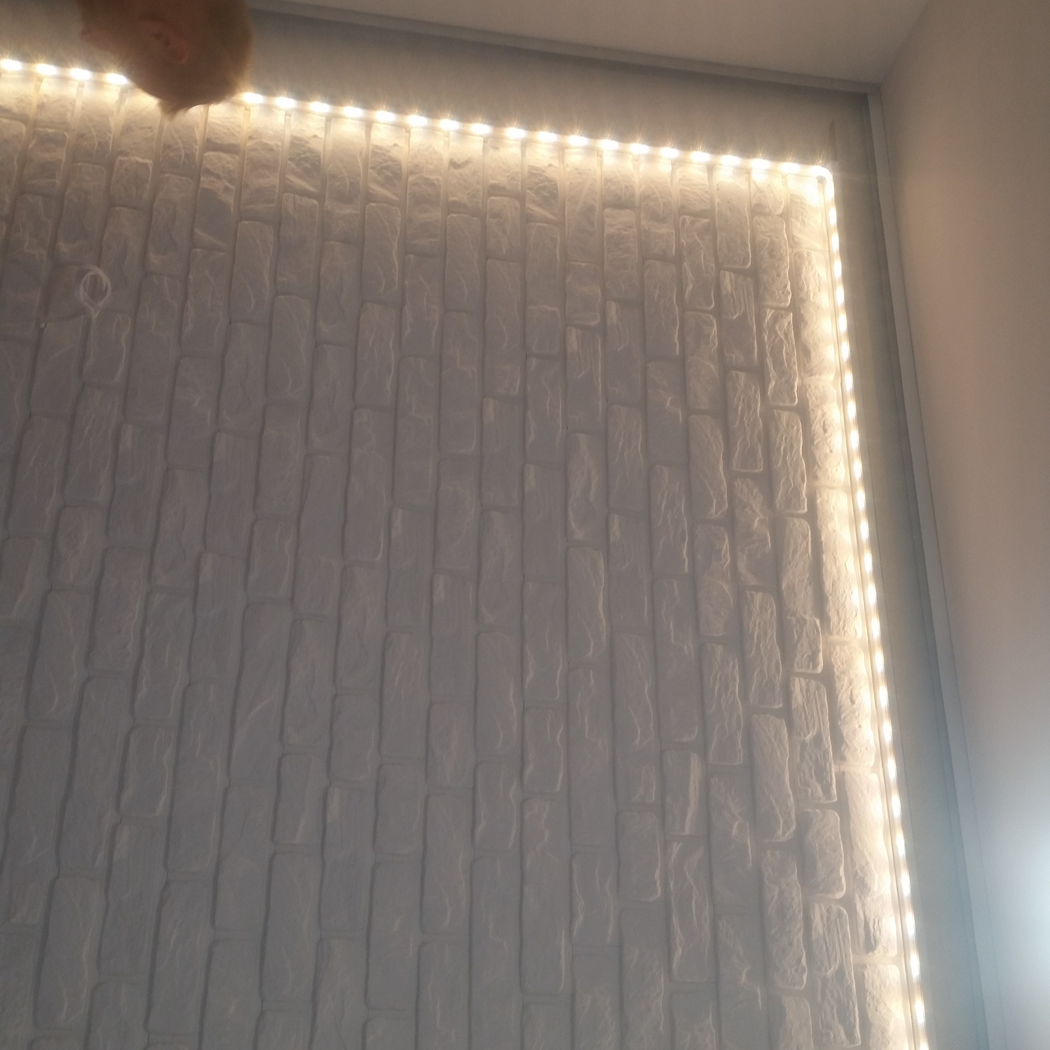 Ściana z białej cegły dekoracyjnej oświetlona taśmą LED, widoczne niedoskonałości w ułożeniu cegieł i wystający fragment czyjejś głowy w rogu.