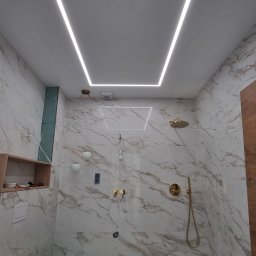 Sufitowski - Nowoczesna łazienka z marmurowymi płytkami, złotą armaturą i podświetlanym sufitem w kształcie prostokąta, widoczne elementy instalacji elektrycznej.