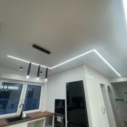 Nowoczesna kuchnia z białymi szafkami, czarnym sprzętem AGD i oświetleniem LED w suficie, z widoczną lampą wiszącą z czarnymi kloszami i śladami po remoncie na suficie.