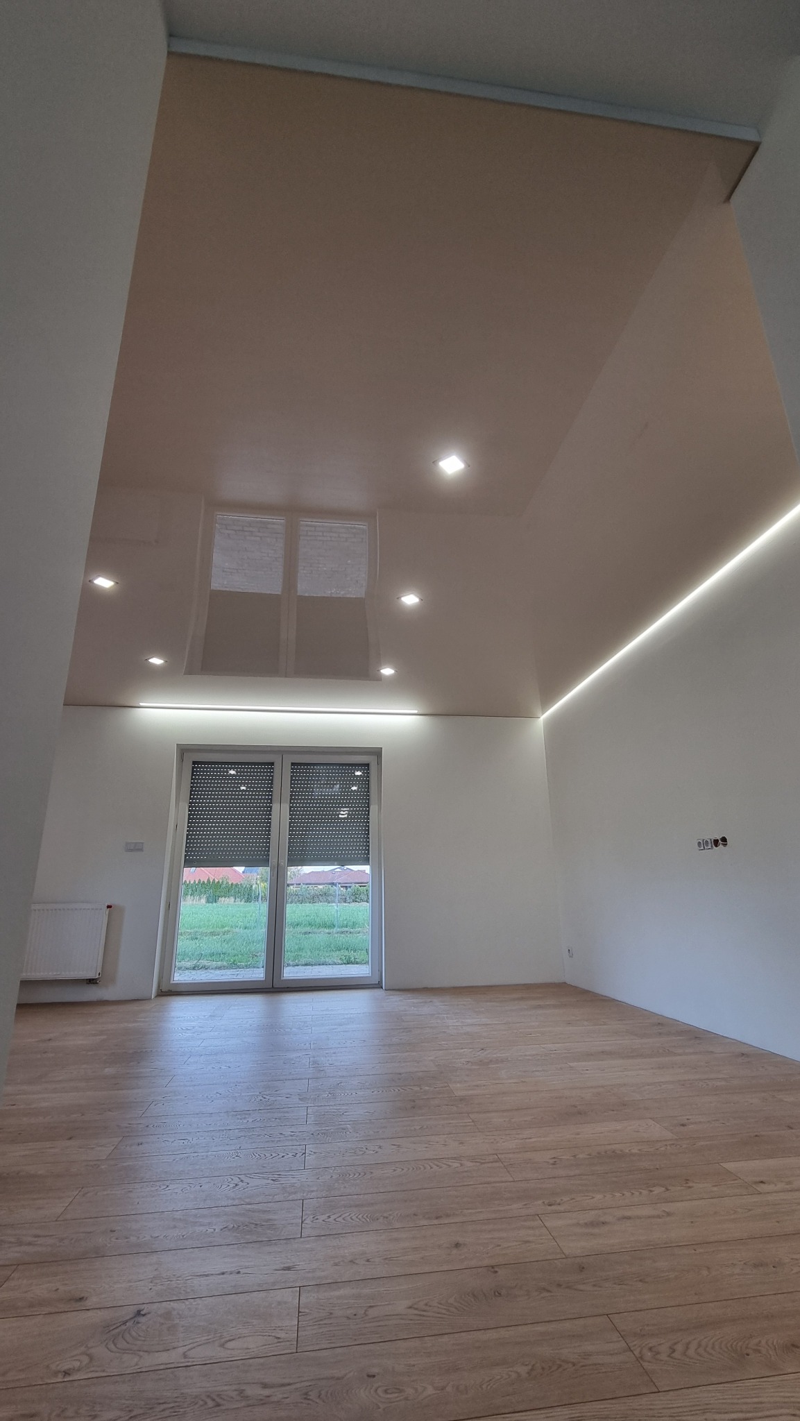 Jasny, przestronny pokój z beżowym, błyszczącym sufitem z wbudowanymi punktowymi i liniowymi źródłami światła LED, drewniana podłoga i okno z roletami zewnętrznymi.