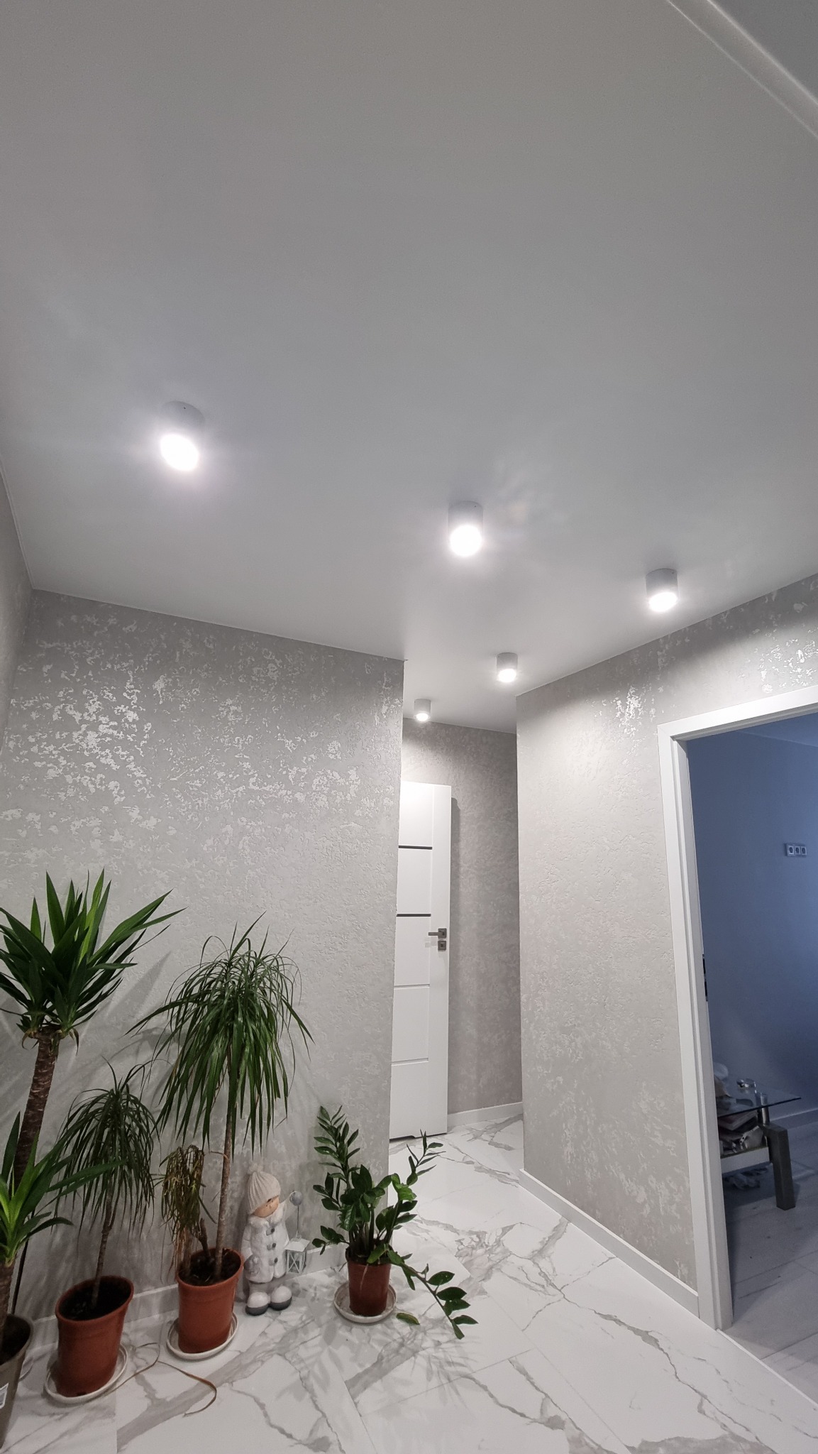 Korytarz z białym, gładkim sufitem z zamontowanymi okrągłymi lampami LED, ściany pokryte szarą tapetą o nieregularnej fakturze, marmurowa podłoga i rośliny doniczkowe wzdłuż ściany.