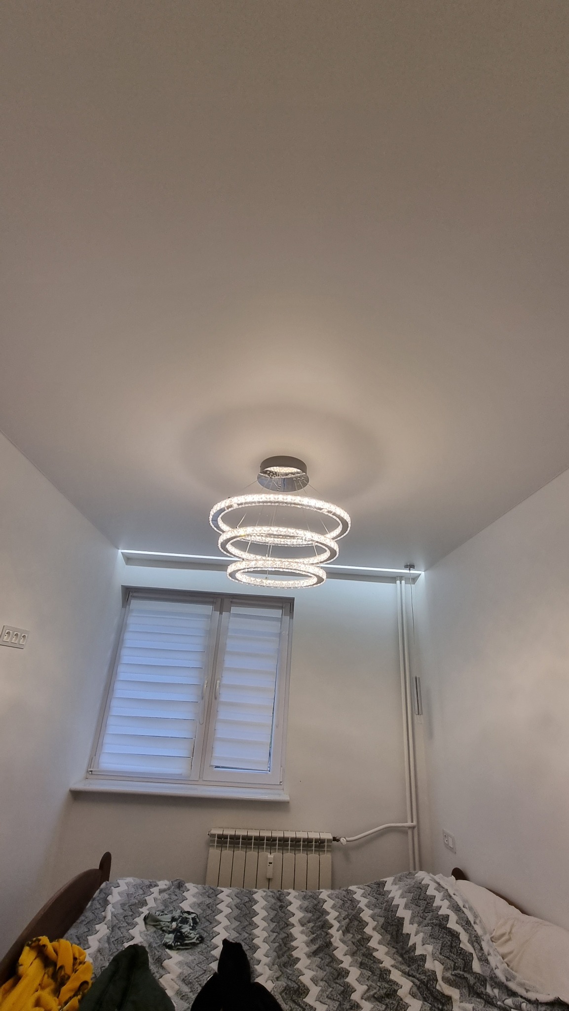 Nowoczesny, trzypoziomowy żyrandol z kryształkami, oświetlający biały sufit w sypialni z grzejnikiem pod oknem.