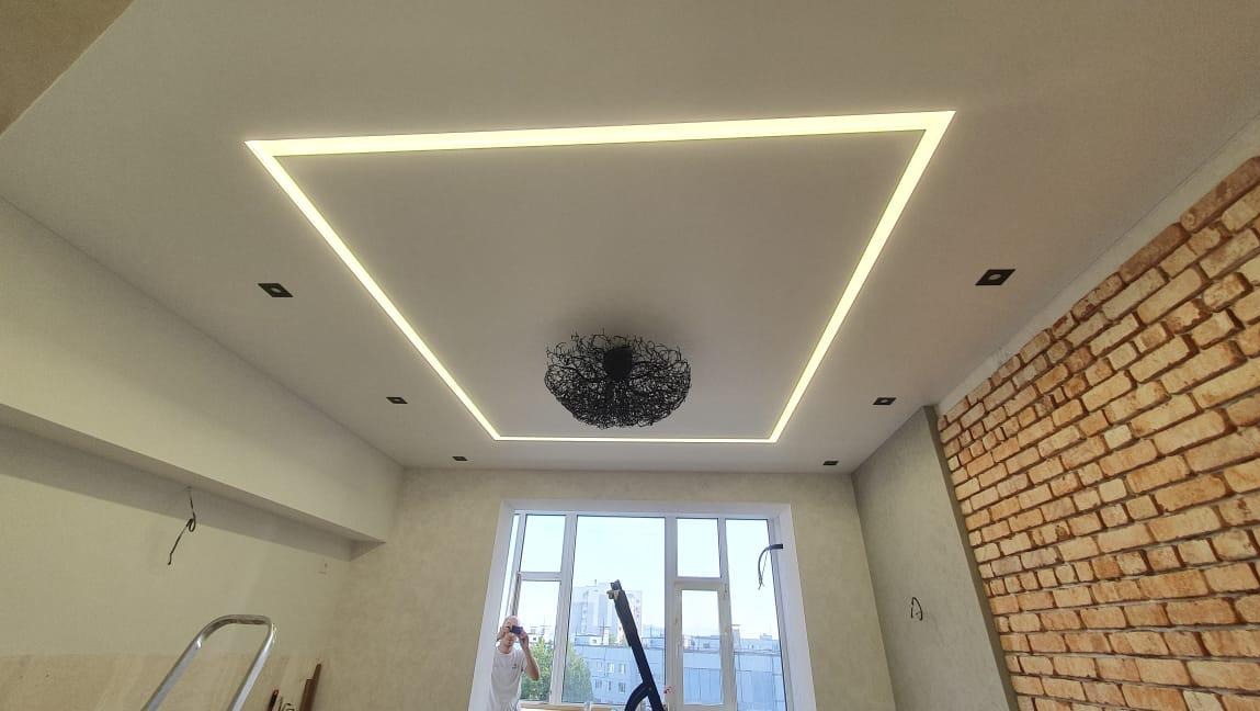 Nowoczesny sufit z podświetleniem LED w kształcie kwadratu, centralnie umieszczony ozdobny żyrandol oraz fragment ściany z cegły dekoracyjnej.