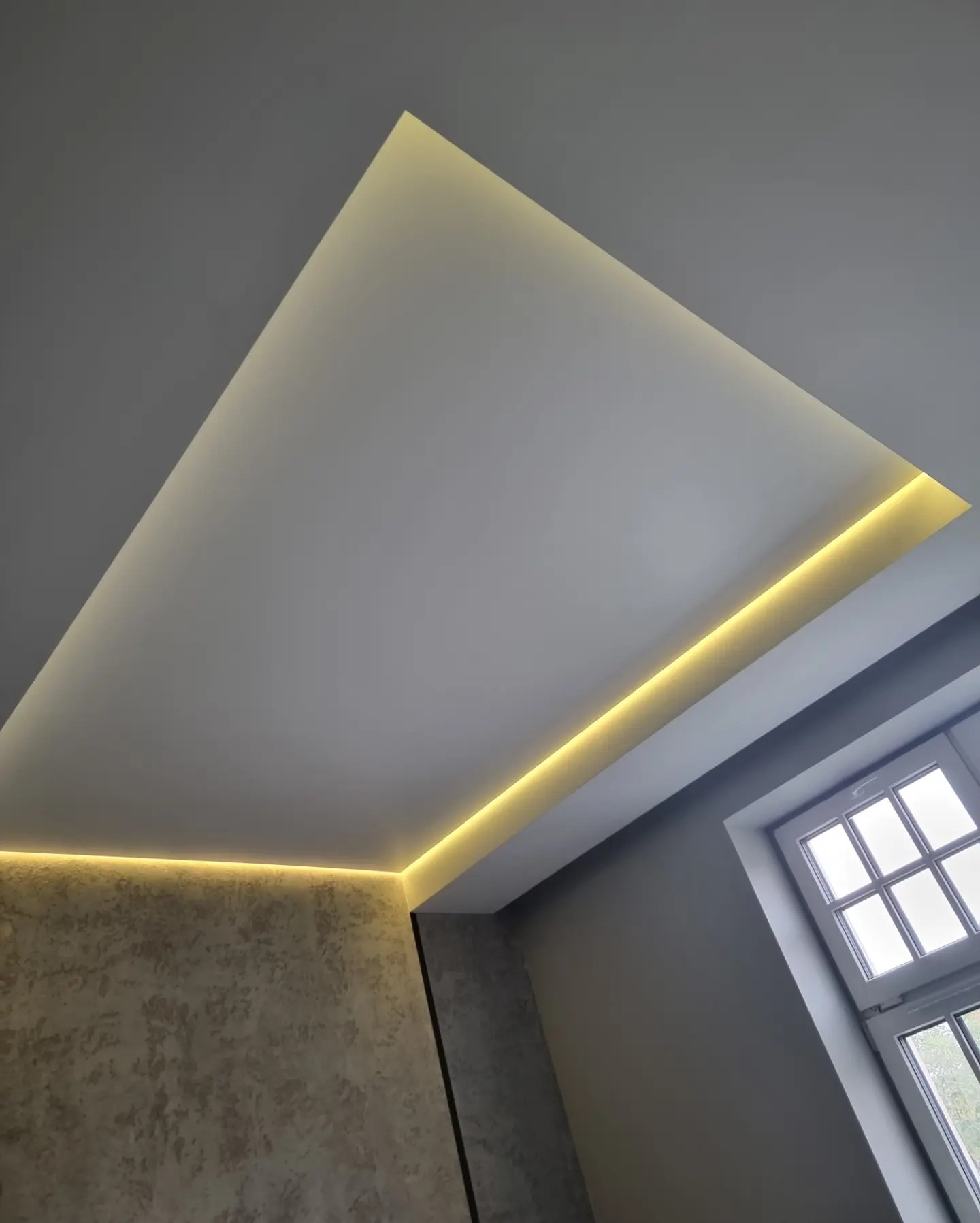 Nowoczesny sufit napinany z oświetleniem LED wzdłuż krawędzi, widok z dołu, okno w prawym rogu, ściana z dekoracyjnym tynkiem.