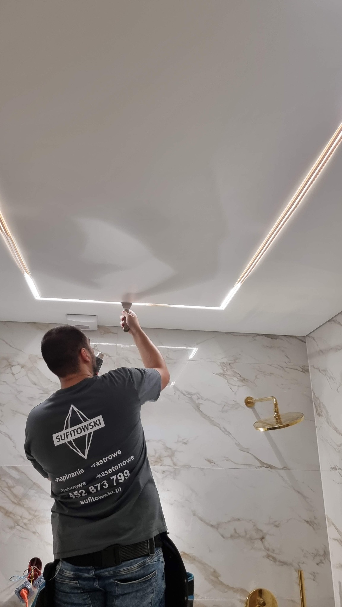 Montaż sufitu z oświetleniem LED w łazience z marmurowymi płytkami; widoczny pracownik z narzędziem i logo firmy na koszulce.