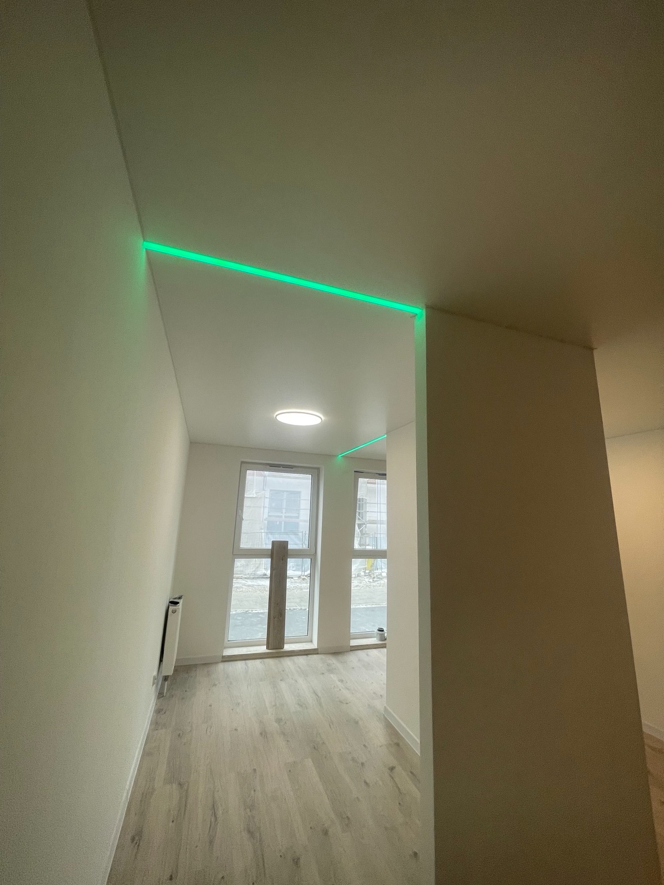 Nowoczesne wnętrze z oświetleniem LED wzdłuż krawędzi sufitu, tworzące zieloną linię światła, widoczne okna i fragment podłogi.