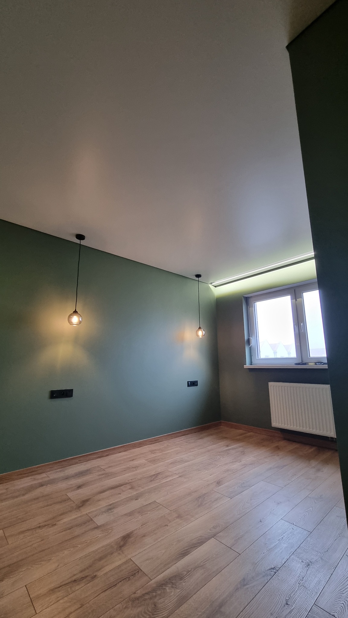 Pokój z zielonymi ścianami, drewnianą podłogą, białym sufitem z ukrytym oświetleniem LED, dwoma wiszącymi lampami z żarówkami w stylu retro i grzejnikiem pod oknem.