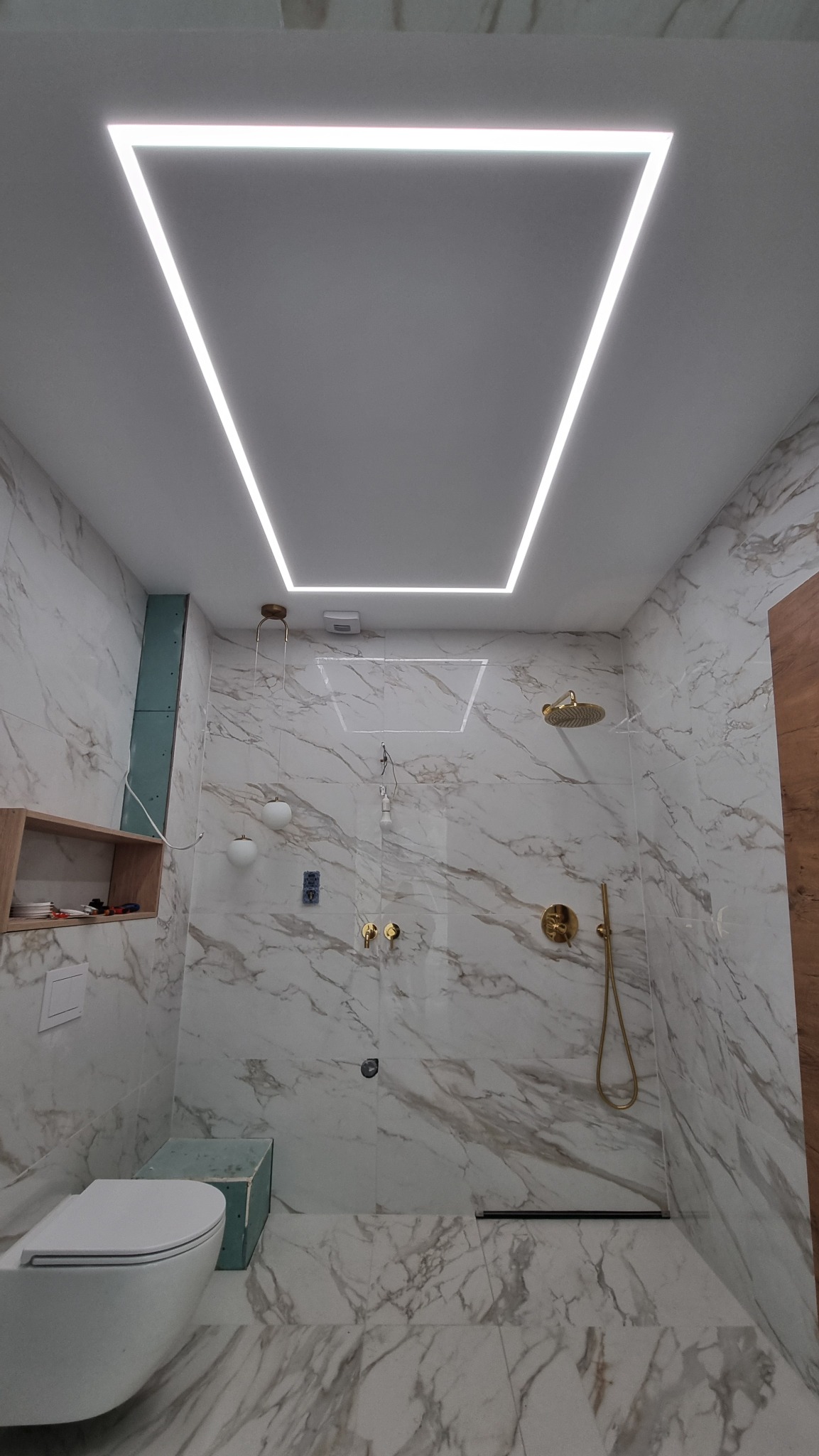 Nowoczesna łazienka z marmurowymi płytkami, złotą armaturą i podświetlanym sufitem w kształcie prostokąta, widoczne elementy instalacji elektrycznej.