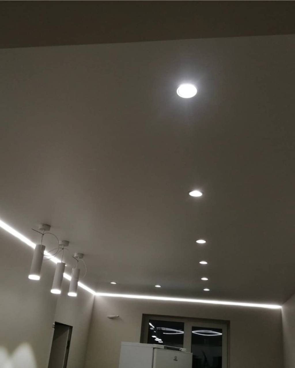 Nowoczesny sufit z oświetleniem LED liniowym i punktowym, uzupełniony wiszącą lampą z cylindrycznymi kloszami, widok z dołu.