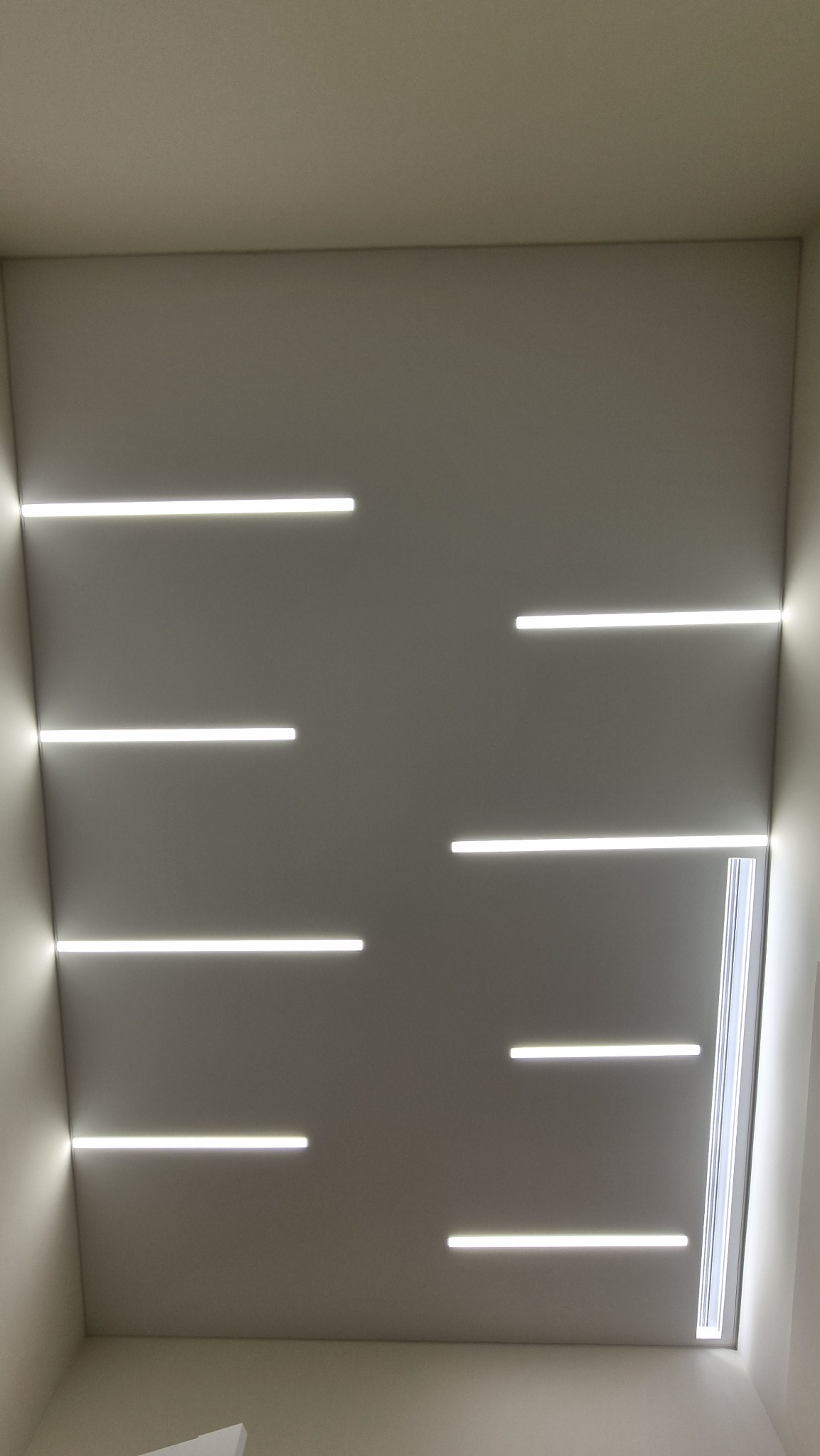 Nowoczesny sufit z wbudowanymi, poziomymi paskami LED, rozmieszczonymi asymetrycznie, z subtelnym oświetleniem krawędziowym i widocznym fragmentem okna dachowego w prawym rogu.