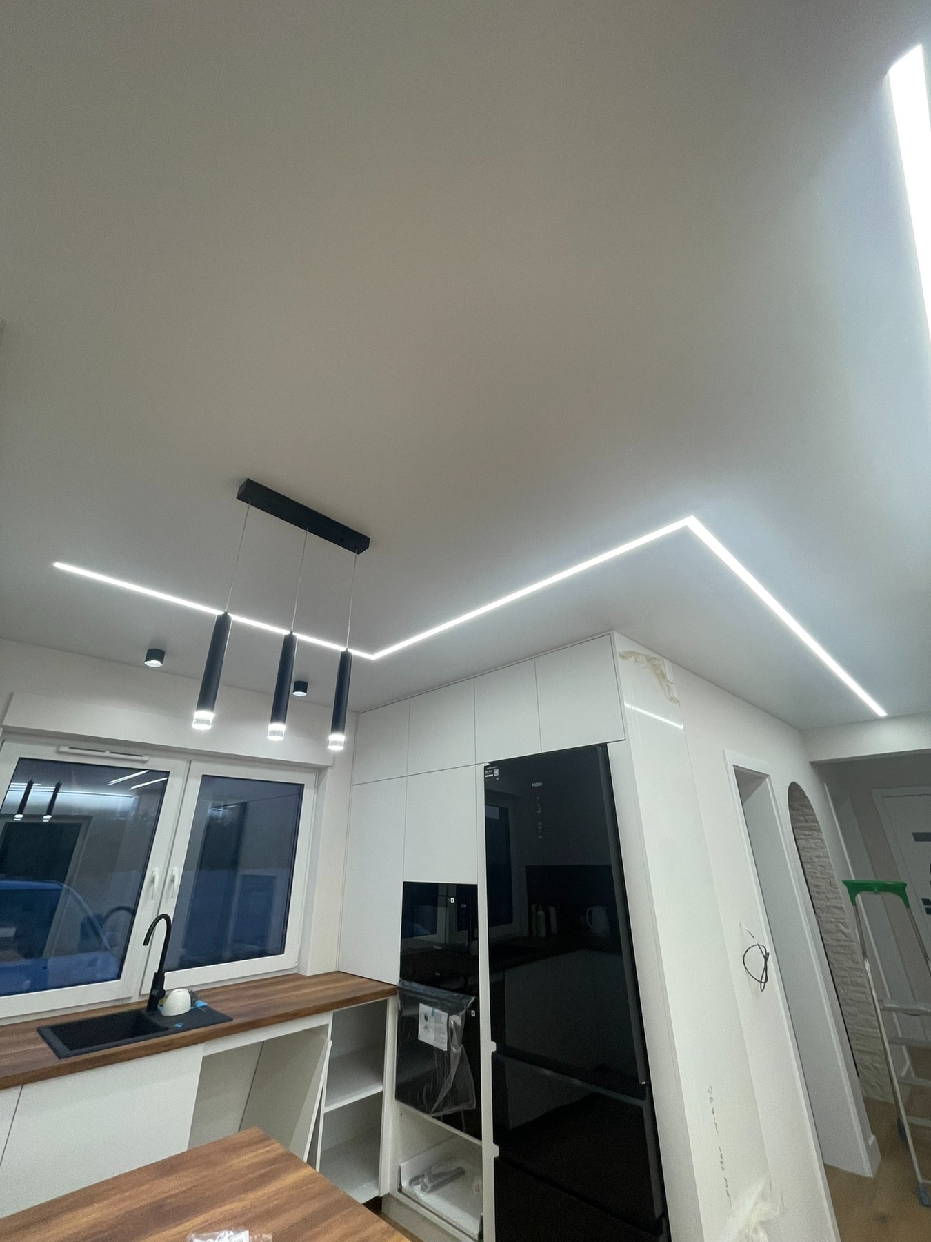 Nowoczesna kuchnia z białymi szafkami, czarnym sprzętem AGD i oświetleniem LED w suficie, z widoczną lampą wiszącą z czarnymi kloszami i śladami po remoncie na suficie.