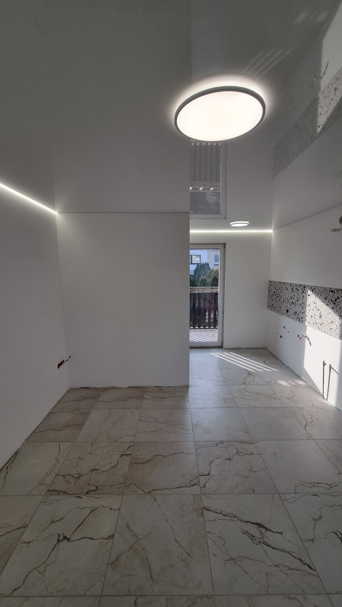 Nowoczesne wnętrze z jasnymi, gładkimi ścianami, marmurową podłogą i oświetleniem LED. Okrągła lampa sufitowa i listwy LED podkreślają minimalistyczny design.