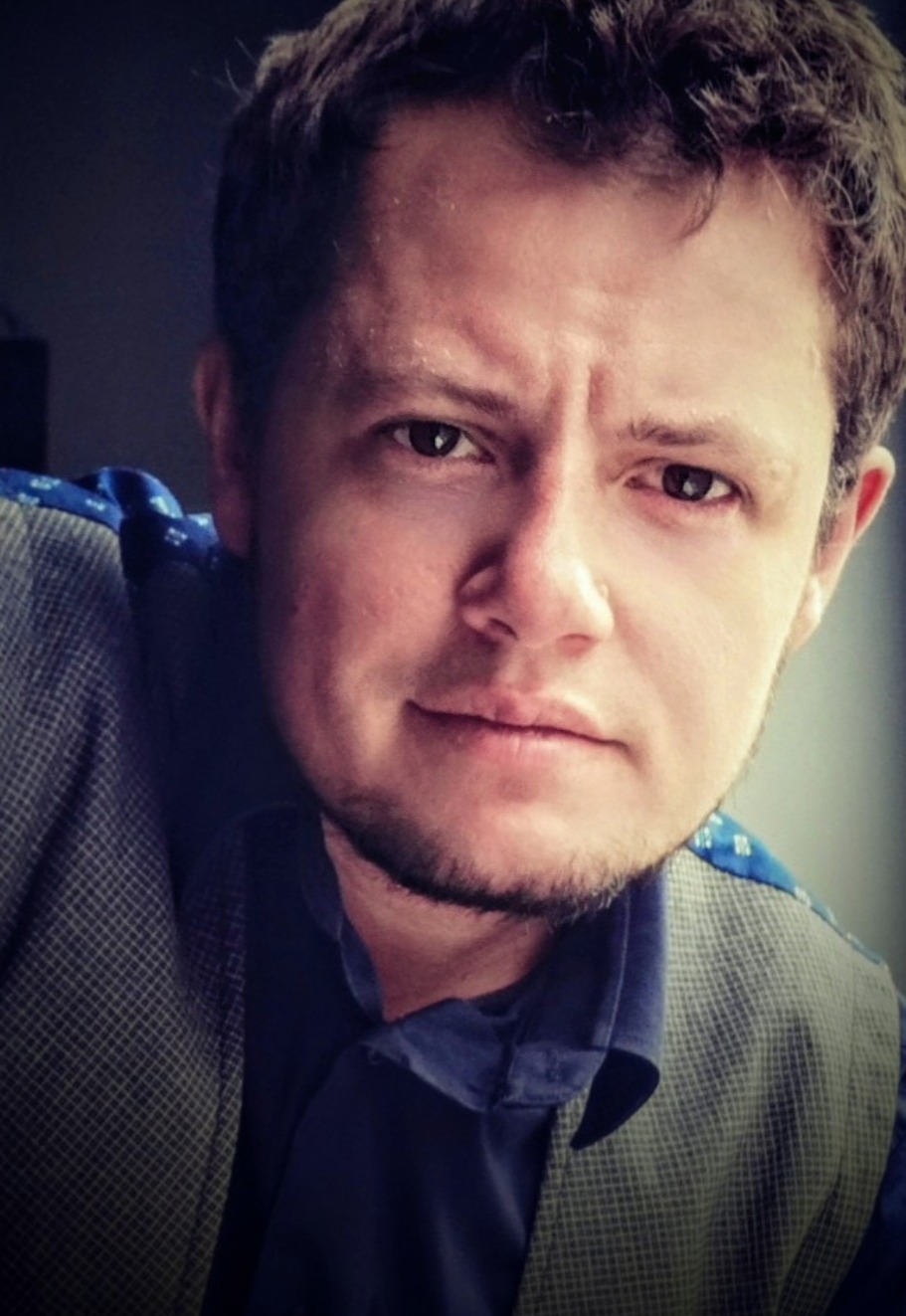 Portret mężczyzny w koszuli i kamizelce, z lekkim uśmiechem i zmarszczonym czołem, oświetlony z góry, z widocznym zarostem.