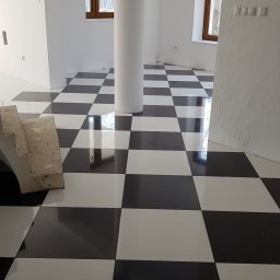 Artur Niziołek - Białe wnętrze z kolumną i błyszczącą podłogą w szachownicę. Widoczne okna z drewnianymi ramami i fragment schodów. Ściana z fakturą.