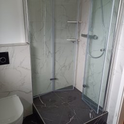 ATN - Kompletna łazienka z kabiną prysznicową typu walk-in, wykończona płytkami imitującymi marmur. Widoczna toaleta i czarna podłoga z odpływem liniowym.
