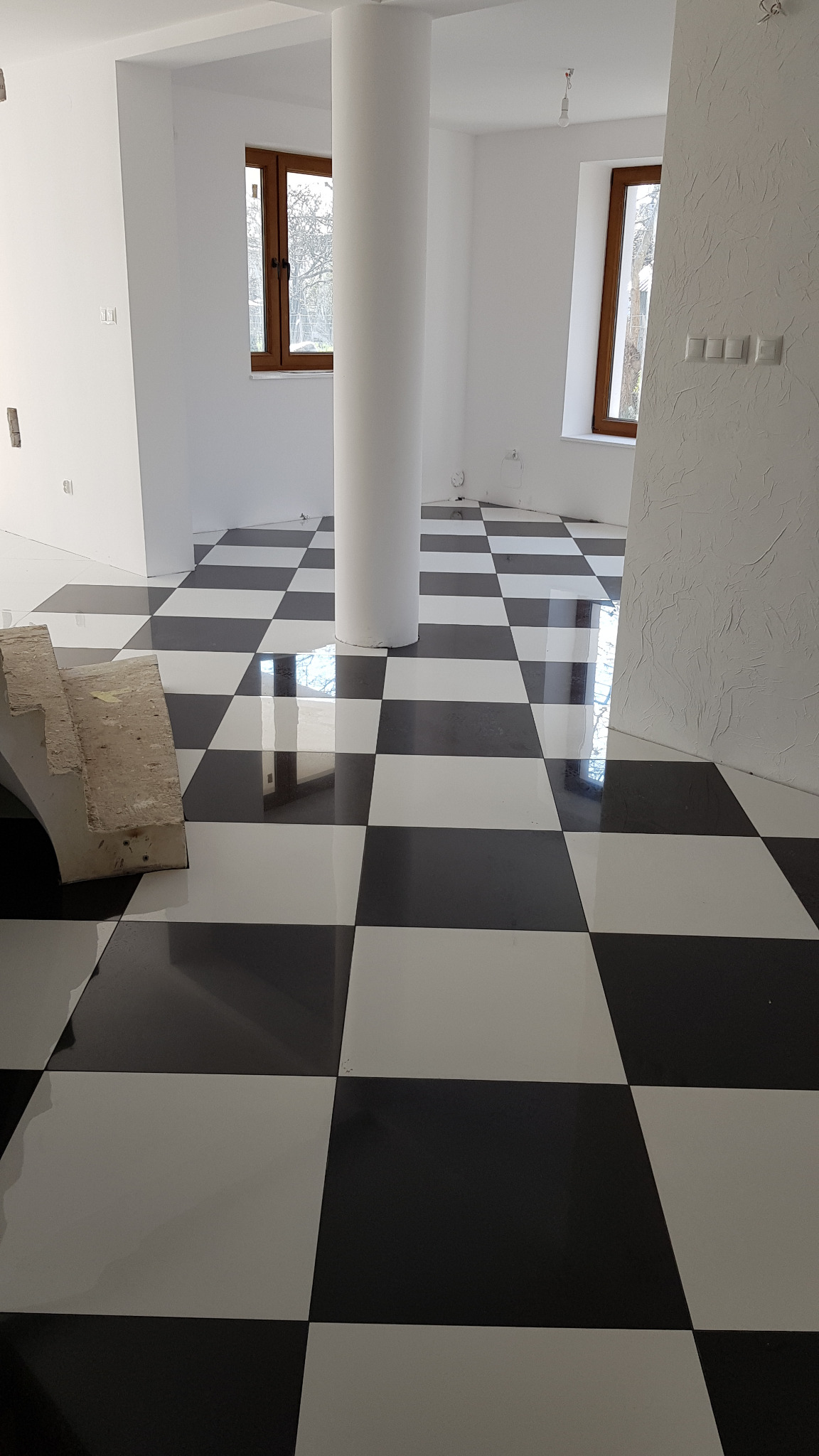 Białe wnętrze z kolumną i błyszczącą podłogą w szachownicę. Widoczne okna z drewnianymi ramami i fragment schodów. Ściana z fakturą.