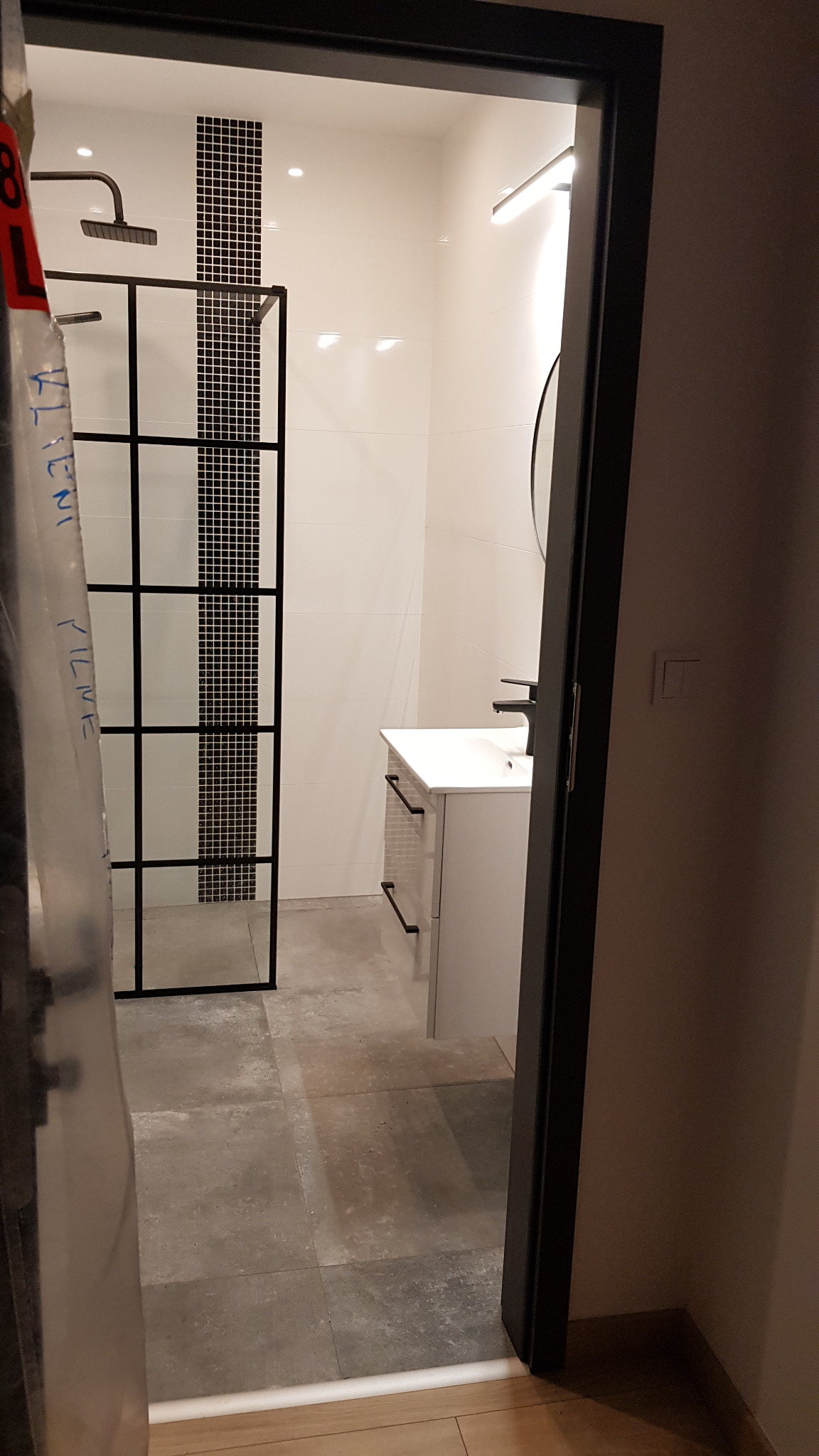 Nowoczesna łazienka z kabiną typu walk-in z czarnymi profilami, białą umywalką z szafką i szarą podłogą z płytek. Widok zza uchylonych drzwi.