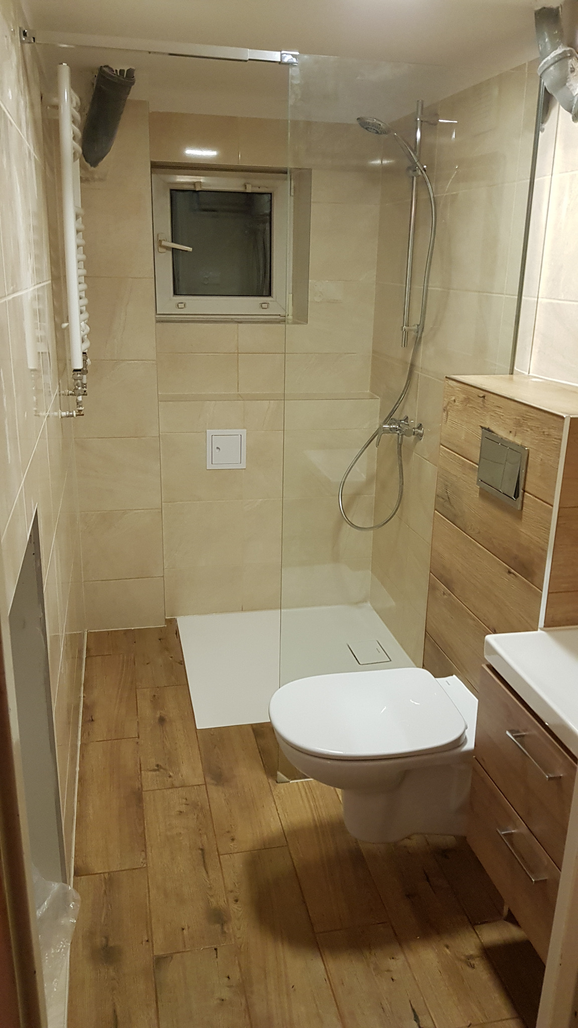 Jasna łazienka z kabiną prysznicową typu walk-in, drewnopodobnymi płytkami na podłodze i ścianach, oraz oknem. Uwagę zwraca drewniana obudowa szafki umywalkowej.