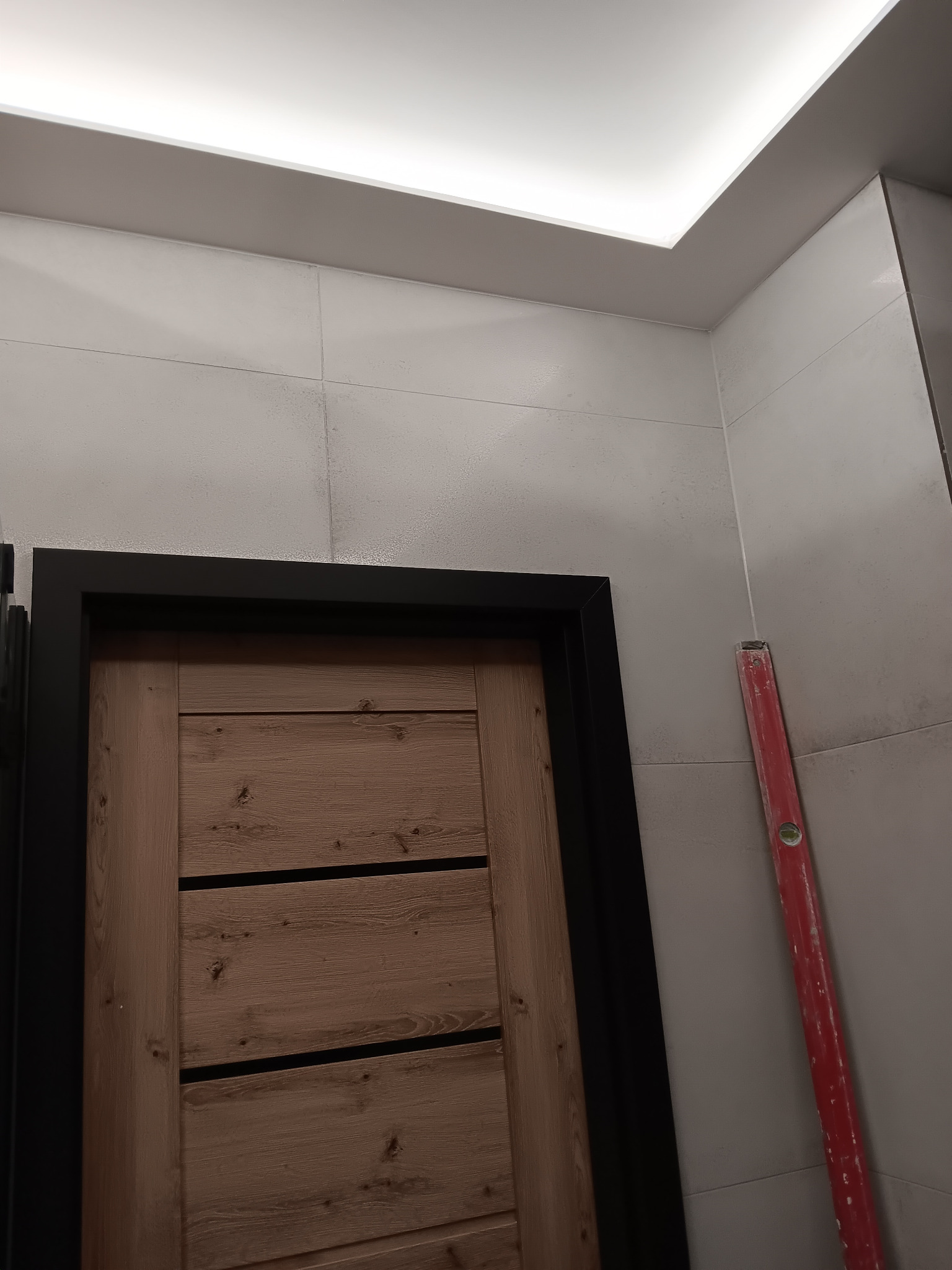 Wykończona łazienka z drewnianymi drzwiami z czarną ramą, szare płytki na ścianach i poziomica oparta o ścianę. Sufit podwieszany z oświetleniem LED.