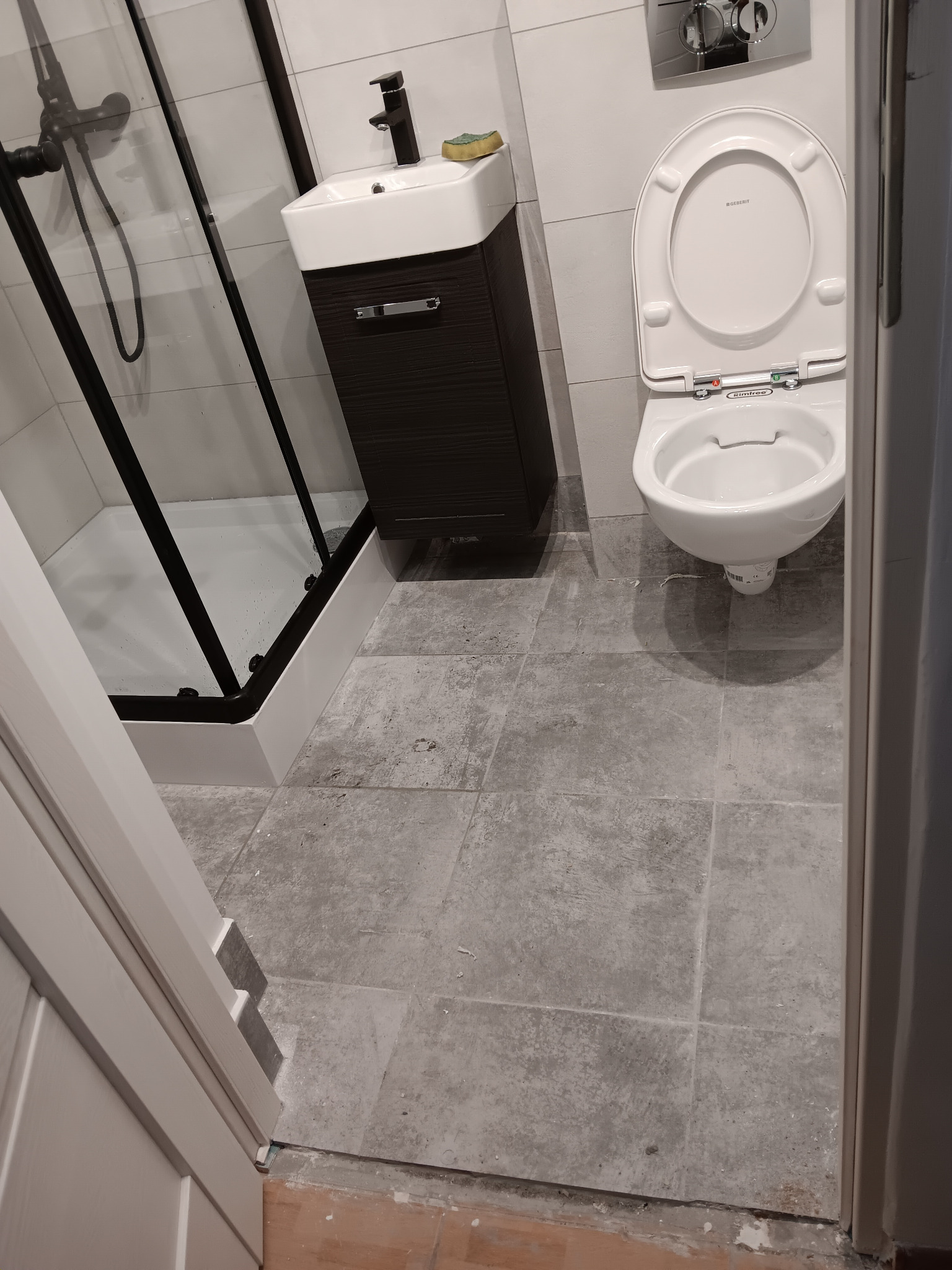 Kompletna łazienka z kabiną prysznicową z czarnymi ramami, umywalką z szafką w kolorze ciemnego drewna i podwieszaną toaletą na szarych płytkach podłogowych.