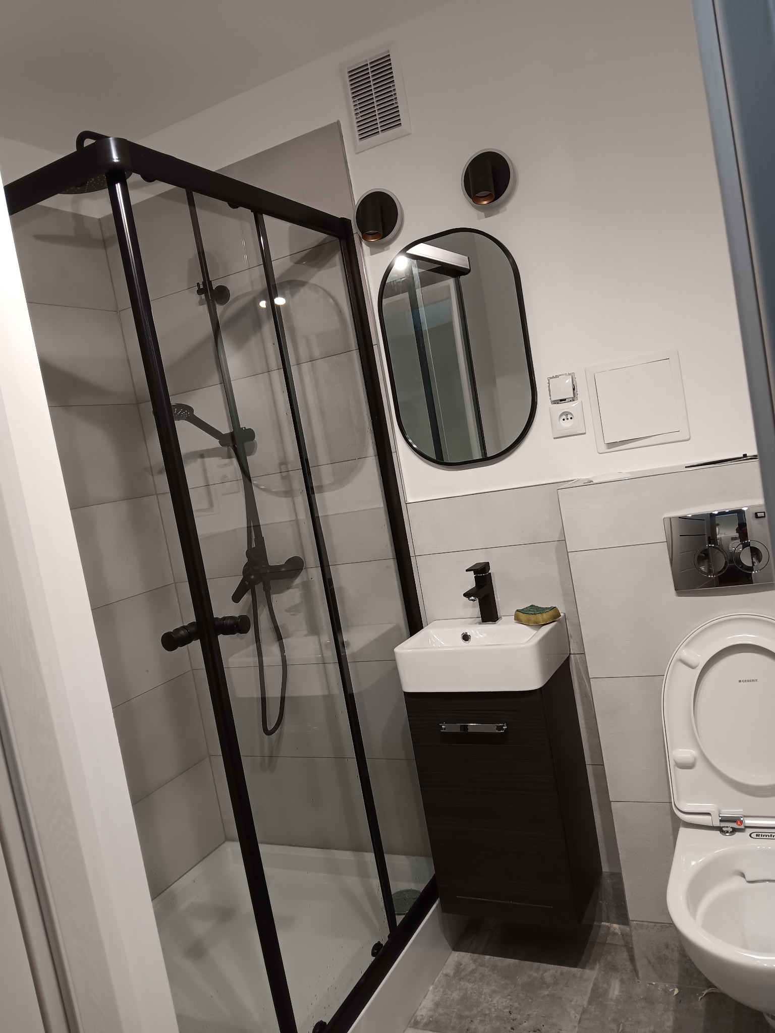 Nowoczesna łazienka z czarną kabiną prysznicową, owalnym lustrem i minimalistyczną umywalką. Szare płytki i biała armatura tworzą spójną całość.