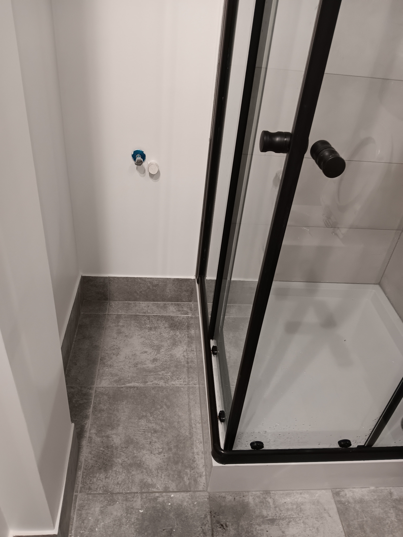 Nowoczesna kabina prysznicowa z czarnymi profilami i szarymi płytkami podłogowymi. Widoczne przyłącza wodne na ścianie. Minimalistyczny design łazienki.