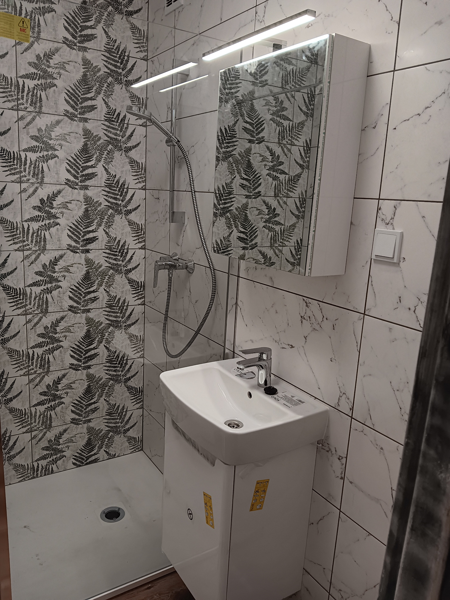 Ukończona łazienka z kabiną prysznicową, umywalką z szafką i lustrem. Ściany wyłożone płytkami imitującymi marmur i płytkami z motywem paproci.