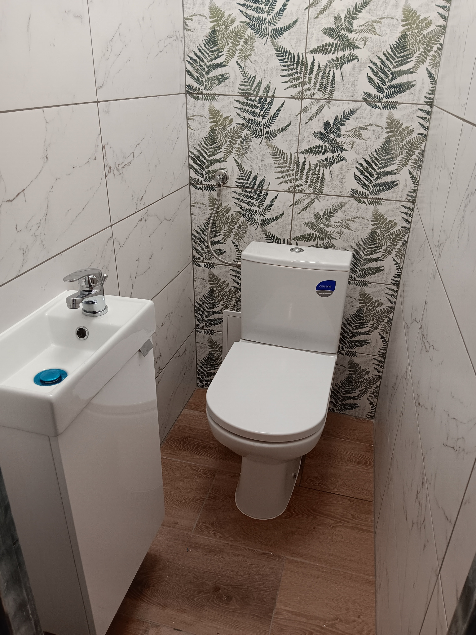 Biała toaleta Cersanit i umywalka z szafką w małej łazience. Ściany wyłożone płytkami imitującymi marmur i płytkami z motywem paproci. Brązowa podłoga.