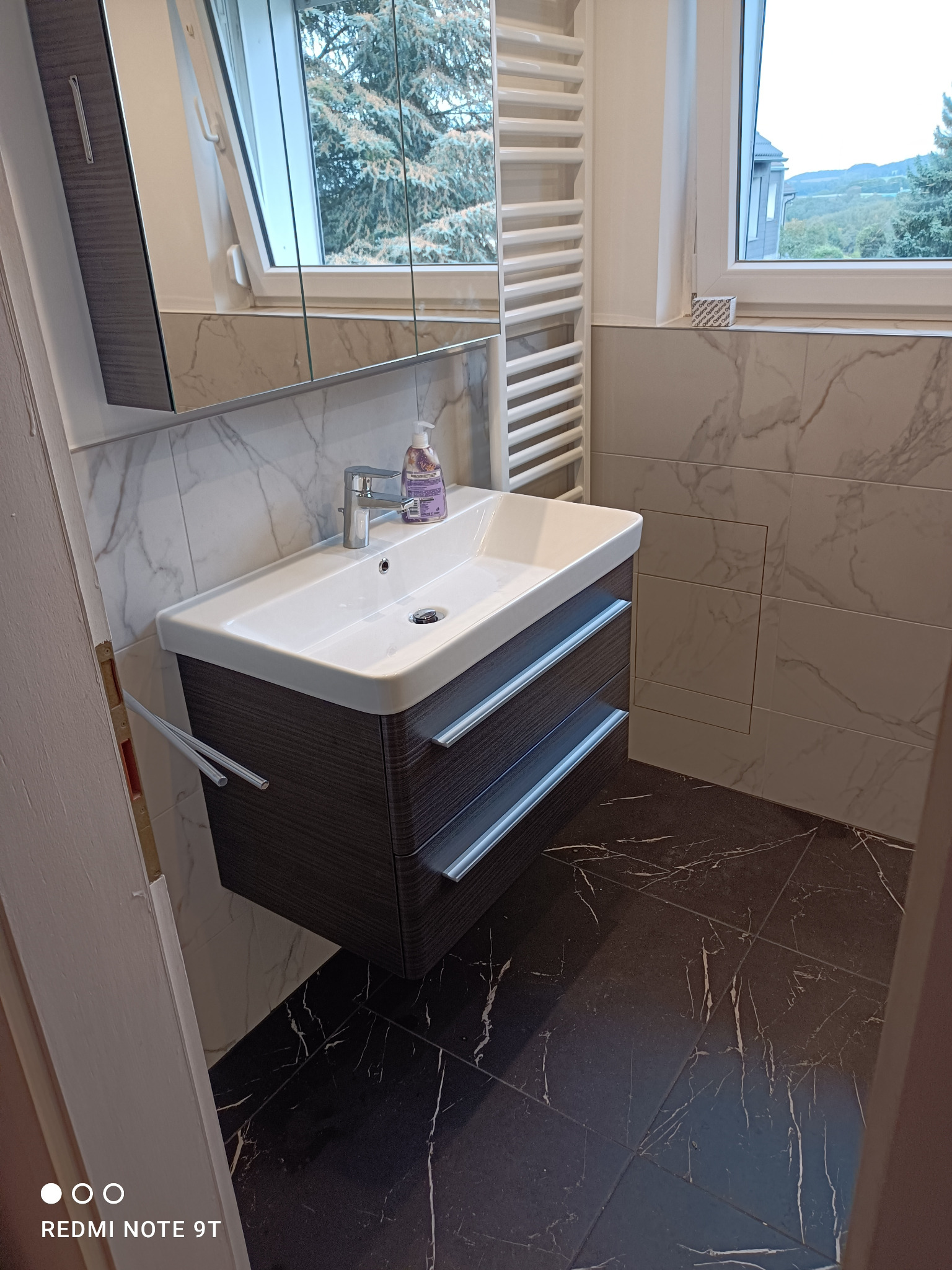 Nowoczesna łazienka po remoncie: biała umywalka z szafką w kolorze ciemnego drewna, marmurowe płytki na ścianach i podłodze, grzejnik drabinkowy, okno z widokiem na zieleń.