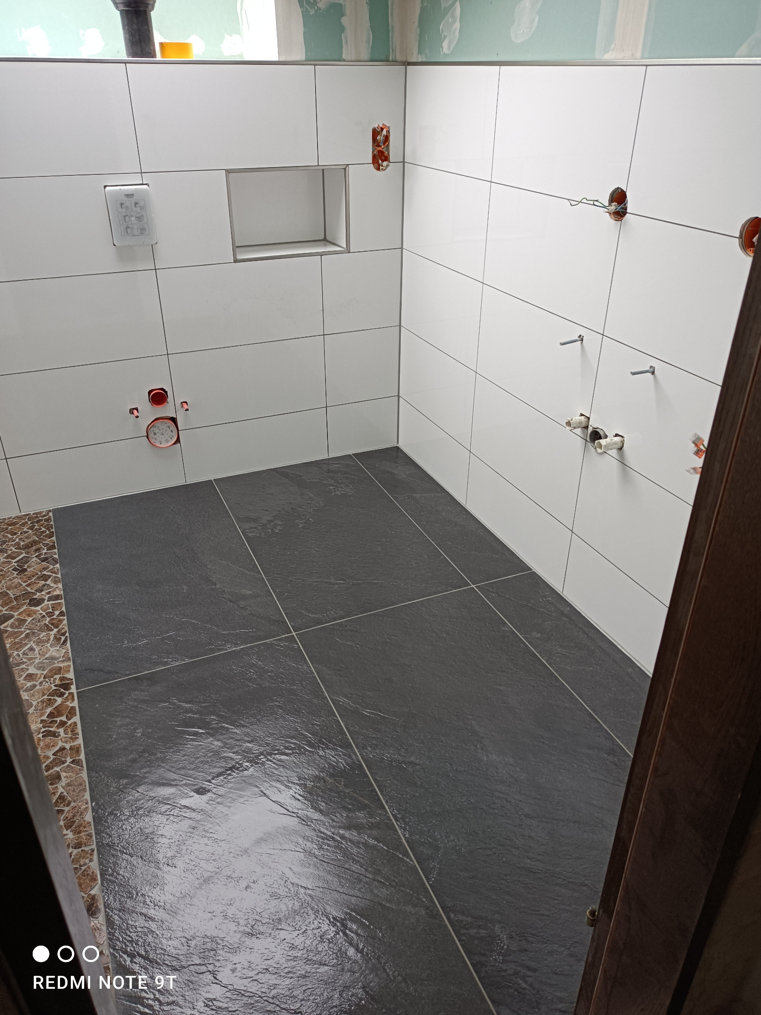 Nowoczesna łazienka w trakcie wykańczania: białe płytki ścienne z wnęką, grafitowa podłoga z płytek, widoczne instalacje wod-kan i elektryczne, dekoracyjny pas mozaiki.