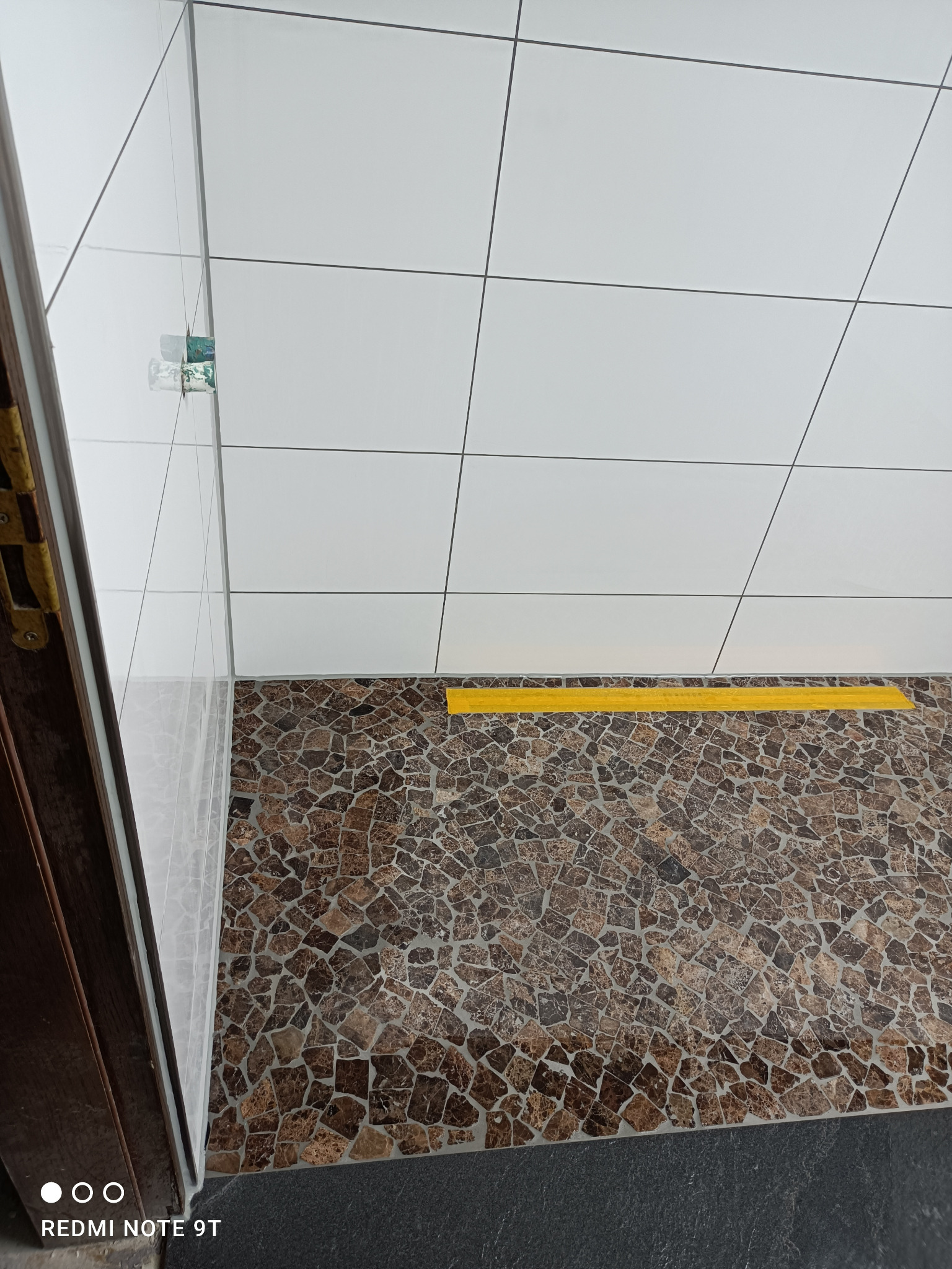 Wykończona łazienka z białymi płytkami na ścianach i mozaikową podłogą w odcieniach brązu, widok z góry, z żółtym akcentem przy podłodze i fragmentem drewnianej futryny.