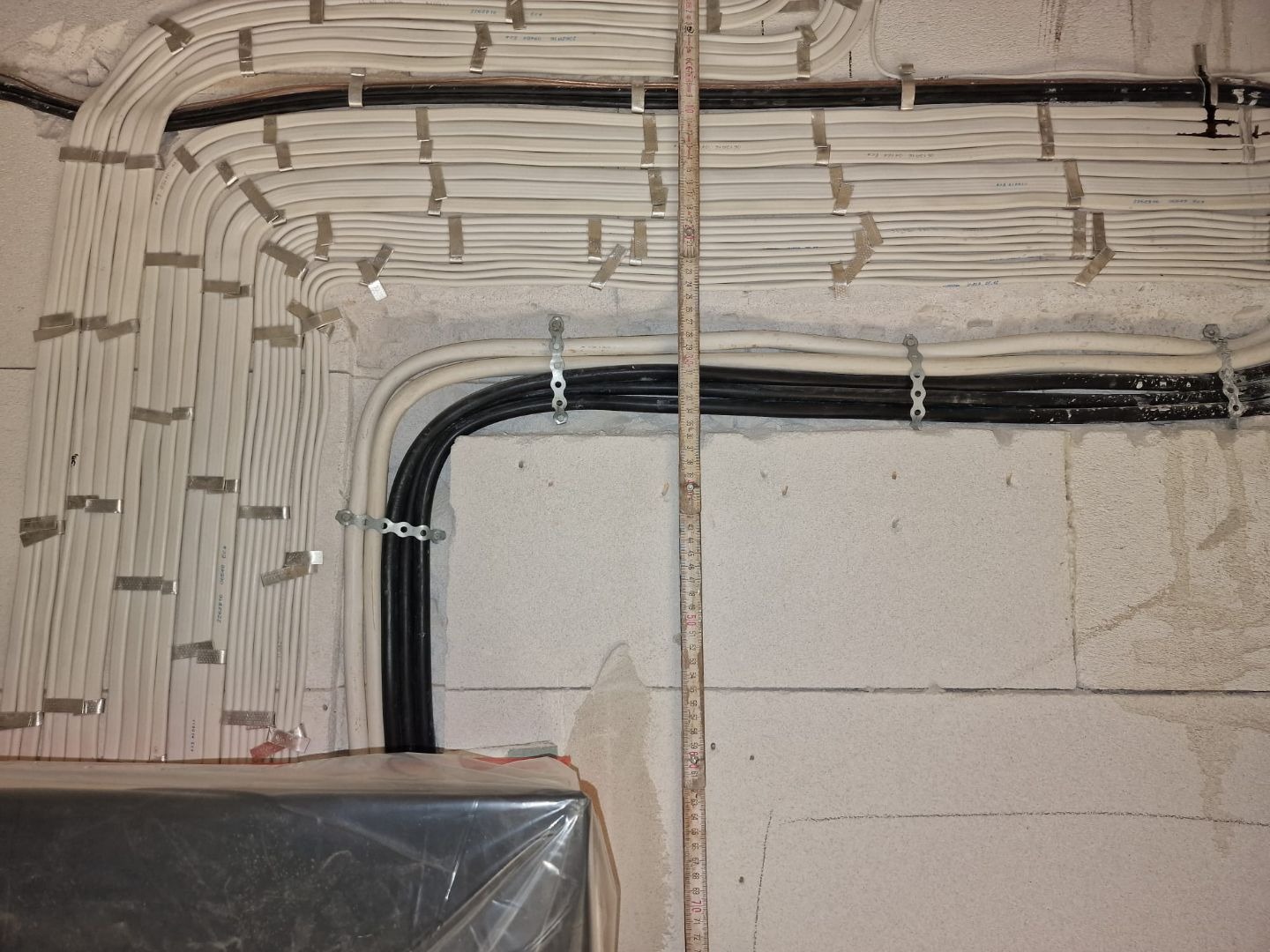 Ściana z surowego betonu z poprowadzonymi wiązkami przewodów elektrycznych w białej i czarnej izolacji, przymocowanych metalowymi uchwytami, obok pionowo umieszczona miarka.