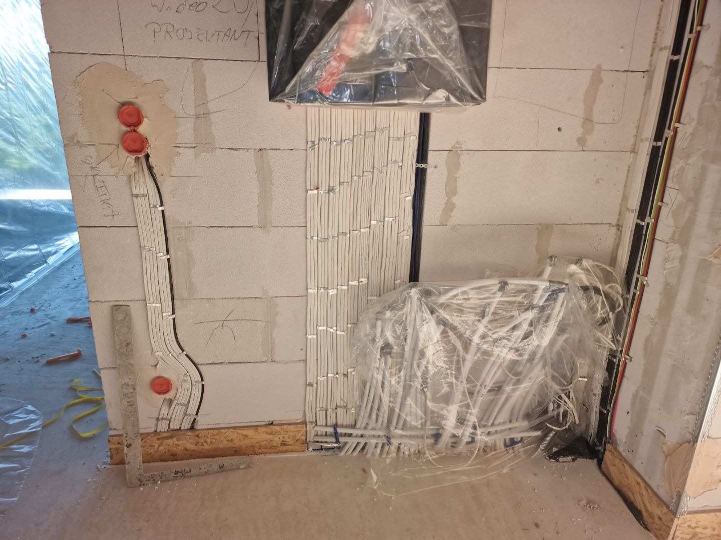 Ściana z betonu komórkowego w trakcie prac elektrycznych: widoczne kable w peszlach ułożone pionowo i poziomo, puszki elektryczne w kolorze pomarańczowym, zwoje kabli zabezpieczone folią,...