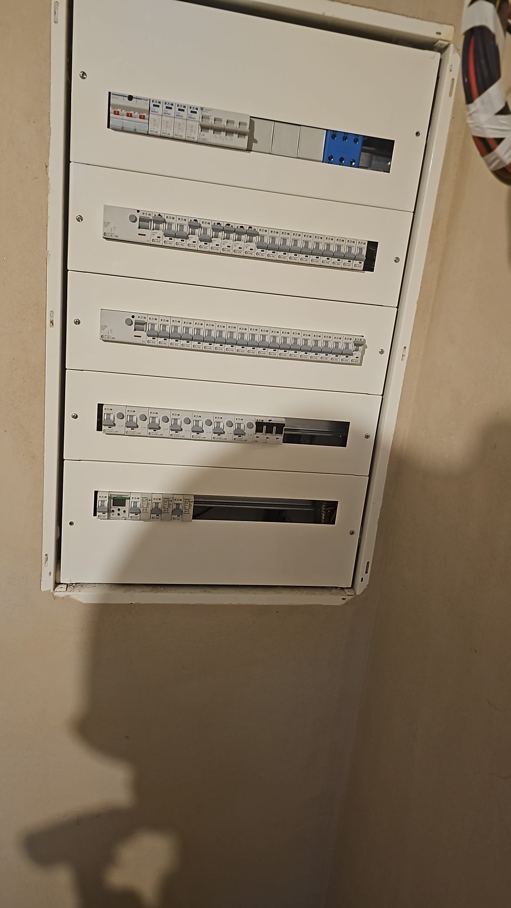 Rozdzielnica elektryczna z wieloma zabezpieczeniami różnego typu, zamontowana na ścianie o szorstkiej fakturze, z widocznymi śladami po pracach instalacyjnych.