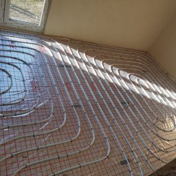 FIRMA SZKOLENIOWO HANDLOWA Zbigniew Adamski - Białe rury ogrzewania podłogowego ułożone na folii z czerwoną siatką, mocowane czarnymi uchwytami, widoczne okno i surowy sufit w tle.