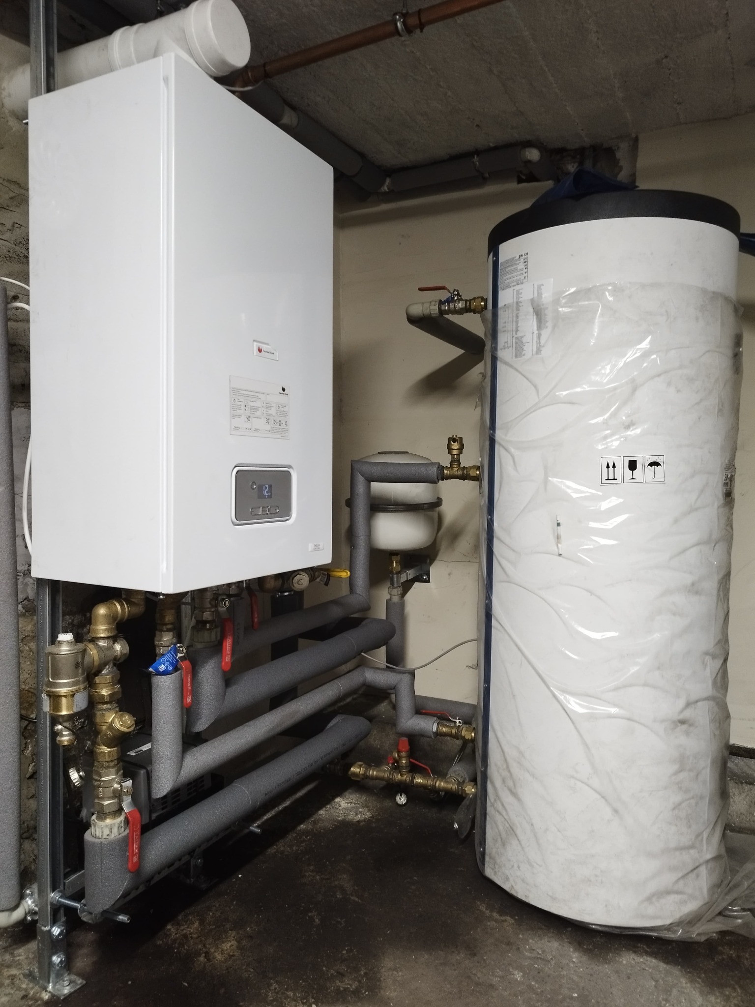Nowy, biały piec gazowy kondensacyjny z podłączonym zasobnikiem ciepłej wody, jeszcze w folii ochronnej, widoczne rury w izolacji termicznej i zawory.