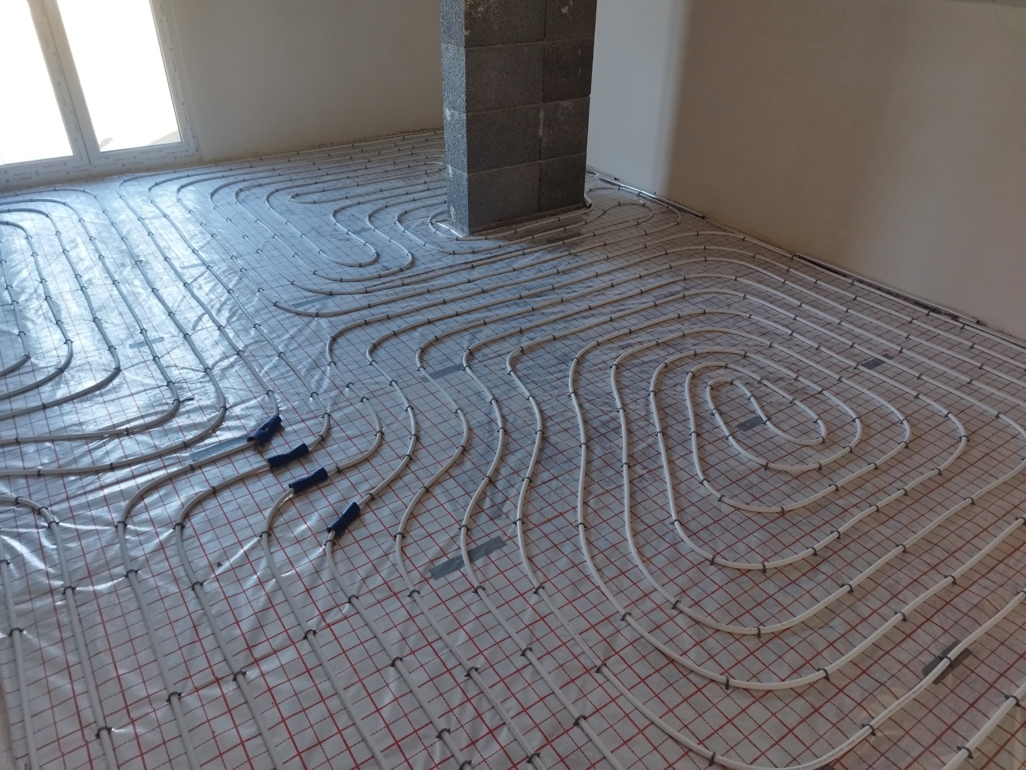 Białe rury ogrzewania podłogowego ułożone spiralnie na folii z siatką w nowym budynku, widoczny betonowy filar i okno w tle.