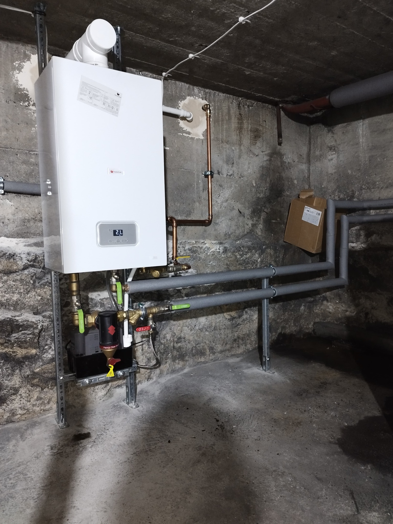 Nowy, biały piec gazowy zamontowany na szarej ścianie z kamienną fakturą, widoczne rury miedziane i szare, kartonowe pudełko w tle.