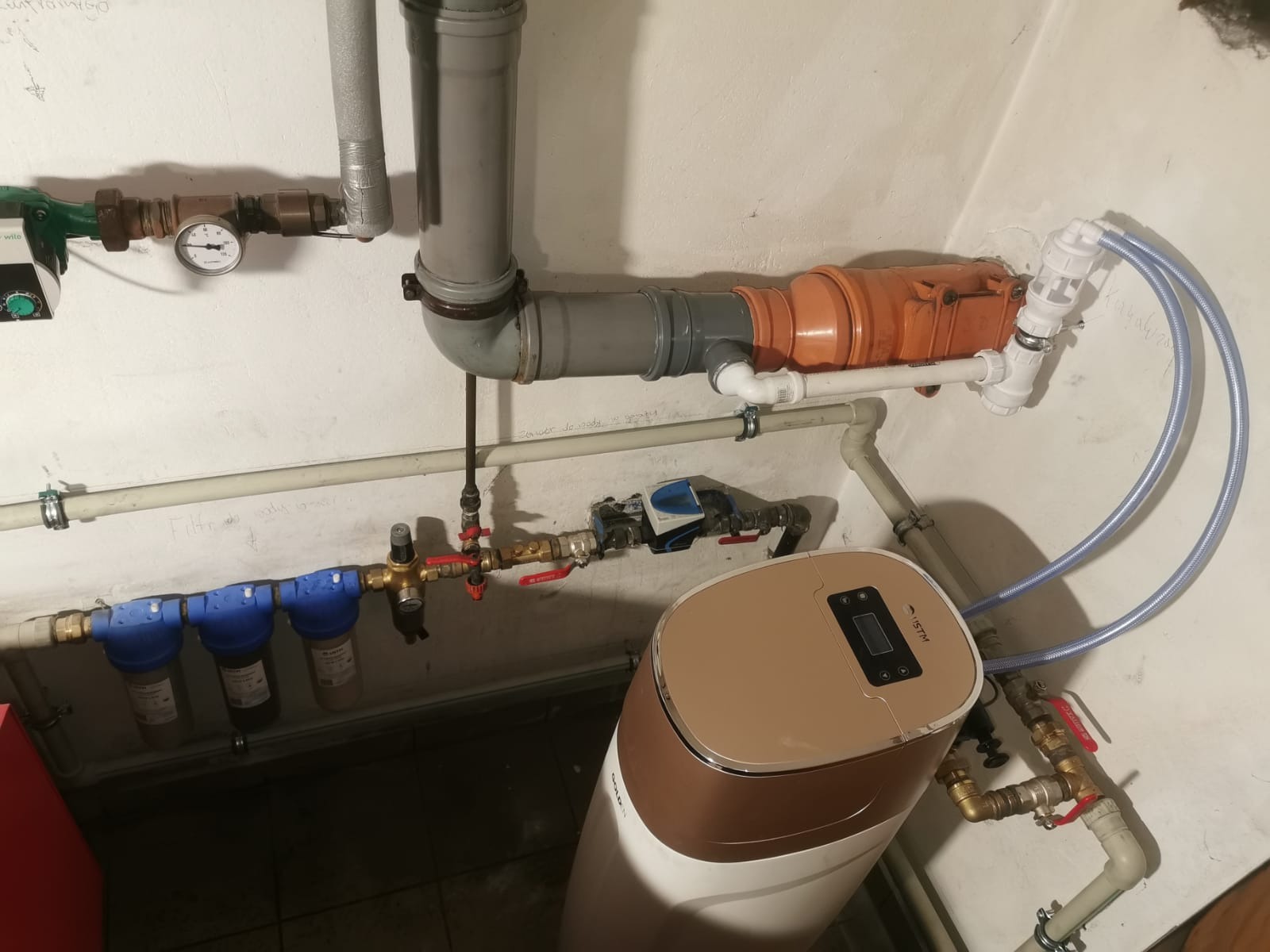 Instalacja wodna z filtrami, rurami i zbiornikiem w kotłowni. Widoczne liczne złącza, zawory i manometr. Ściany pomieszczenia pomalowane na biało.